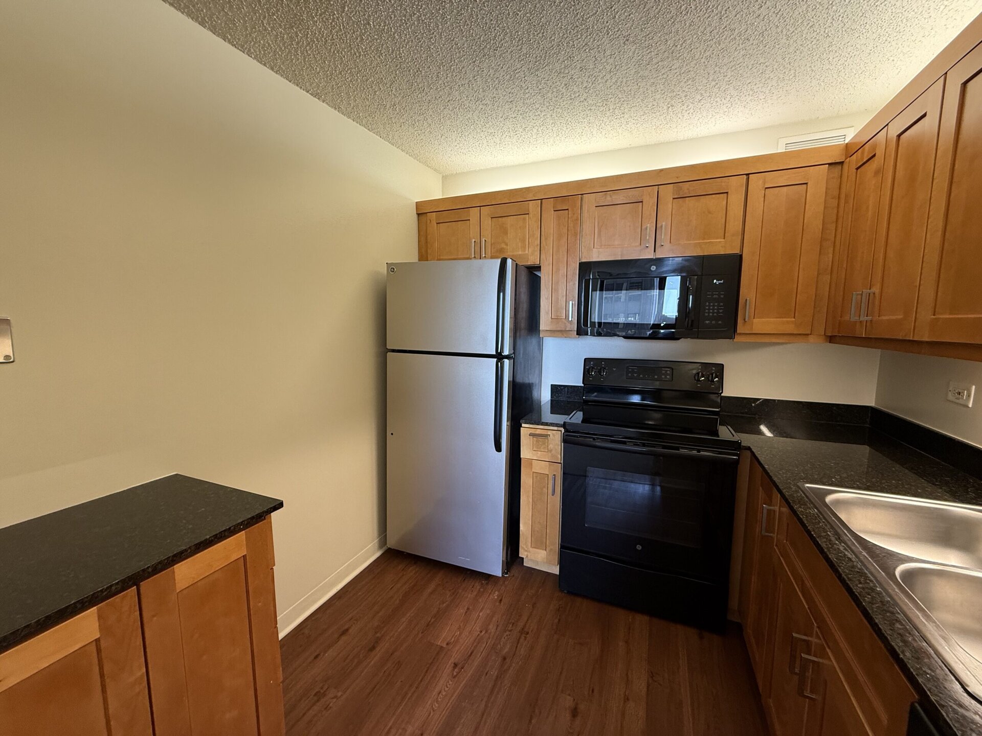 A modern kitchen featuring wooden cabinets with a warm finish, a stainless steel refrigerator, and a black stove and microwave. The countertops are dark granite, and there are dual sinks on the right side. The flooring is a light wood laminate, contributing to a contemporary aesthetic. The walls are painted a soft neutral color, enhancing the overall brightness of the space.