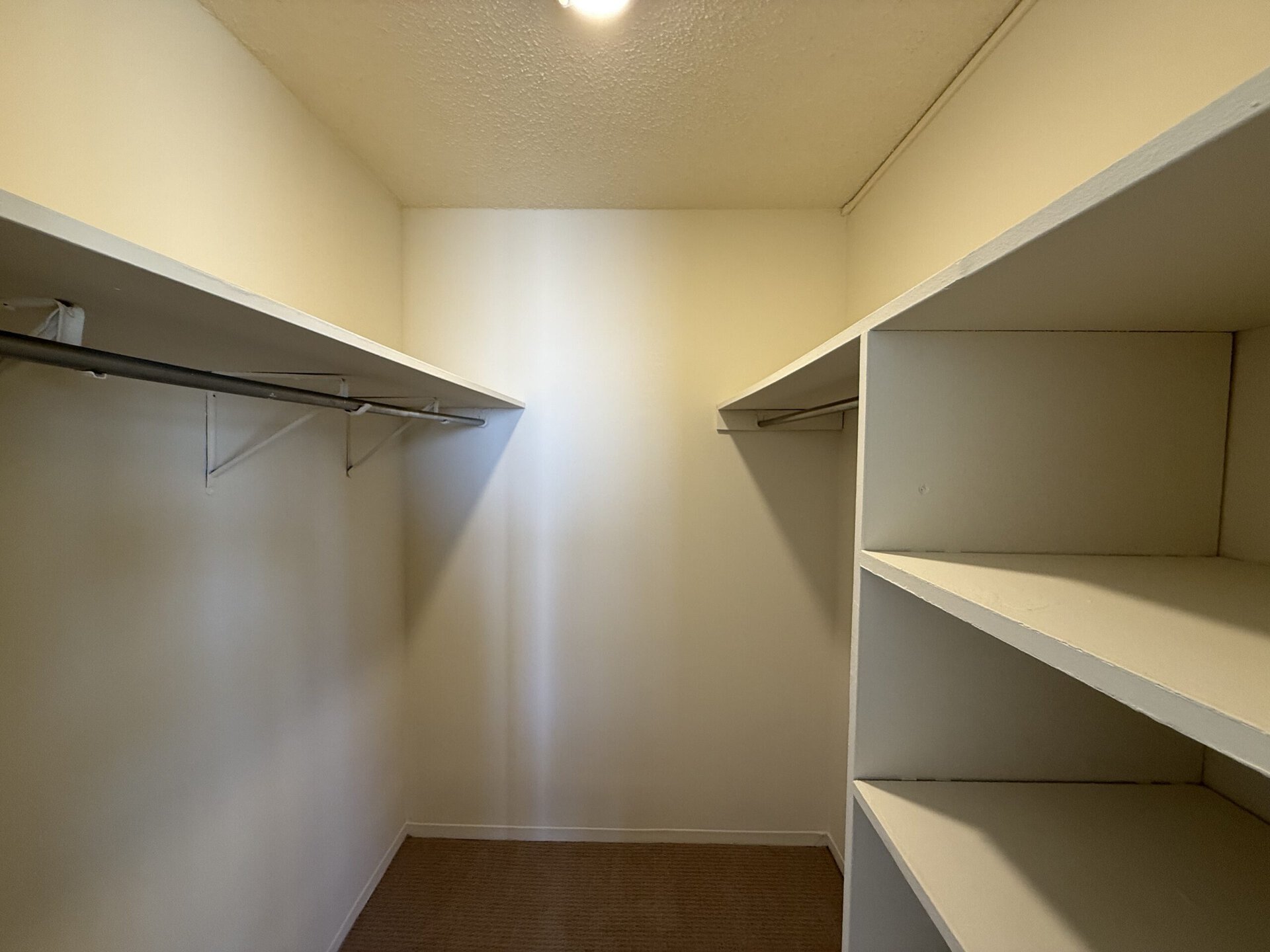 A spacious closet with light-colored walls and a ceiling. On the left side, there is a metal clothing rod supported by brackets, while the right side features open shelves. The overall appearance is clean and uncluttered, providing ample storage space.
