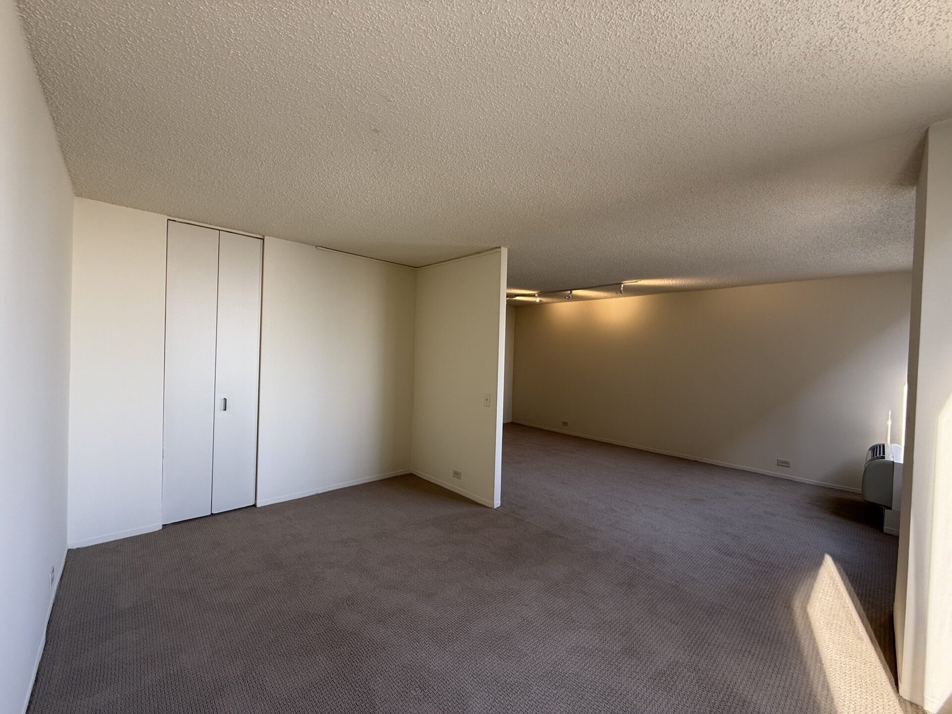 A spacious room with beige carpet and light-colored walls. On the left, there are two closed white closet doors. A partial wall divides the space, leading to a larger area that appears well-lit. The ceiling has a textured finish, and there are track lights installed. In the far corner, an air conditioning unit is visible. The overall atmosphere is bright and open.