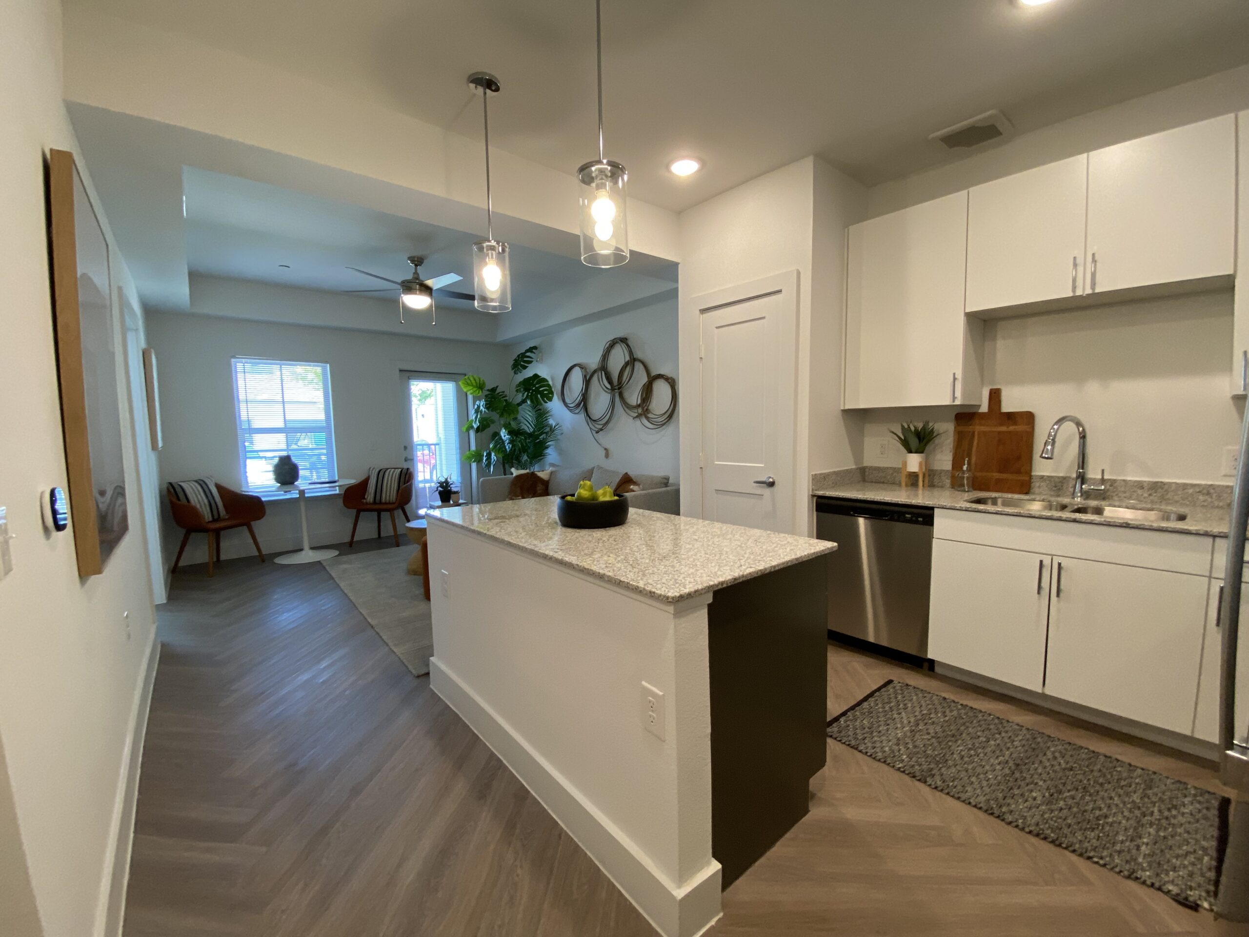 A modern kitchen and living area are visible, featuring a granite countertop island with a bowl of green fruit. The kitchen has white cabinetry, stainless steel appliances, and a wooden cutting board. In the background, a cozy living space includes two orange chairs, a small table, and a large plant. A window allows natural light to enter, and a door leads outside. The flooring is a light wood with a herringbone pattern.