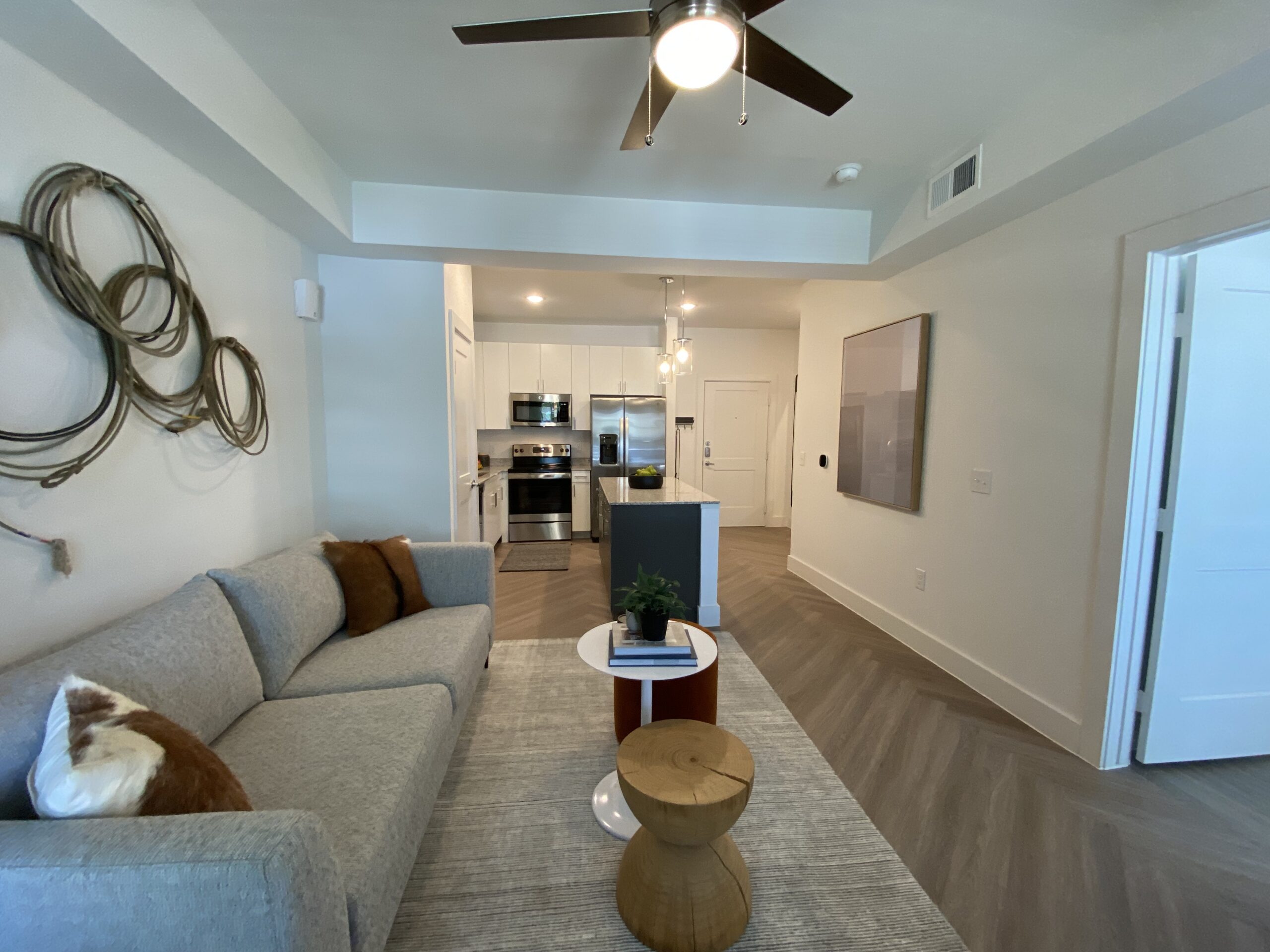 A modern living space features a light gray sofa adorned with decorative pillows, including a brown fur pillow. In front of the sofa, there is a round white coffee table with a stack of books and a small potted plant. The flooring is a herringbone pattern in a light wood tone. To the left, an open kitchen showcases stainless steel appliances, including a refrigerator and an oven, with white cabinetry and a granite countertop island. Above the island, pendant lights hang, adding a contemporary touch. A large piece of abstract art is mounted on the wall, and a door leads to another room.