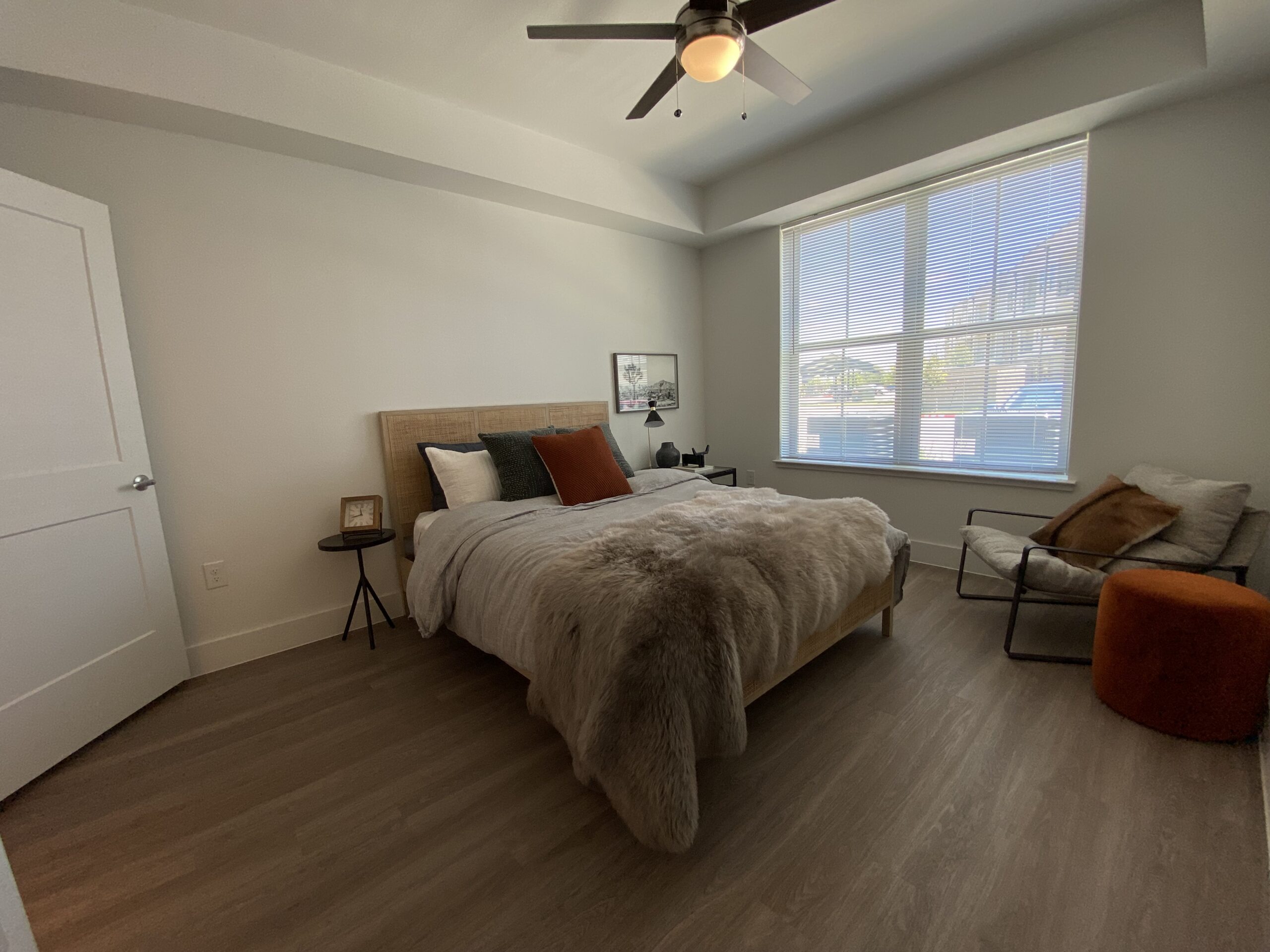 A cozy bedroom features a large bed with a light gray blanket and a plush fur throw. The headboard is made of woven material, and decorative pillows in various colors, including a rust-colored one, add a pop of color. Beside the bed is a small black side table with a clock and a lamp. A comfortable armchair with cushions sits in the corner, next to a round orange ottoman. Natural light streams in through a large window with blinds, illuminating the room's neutral tones and wooden flooring.