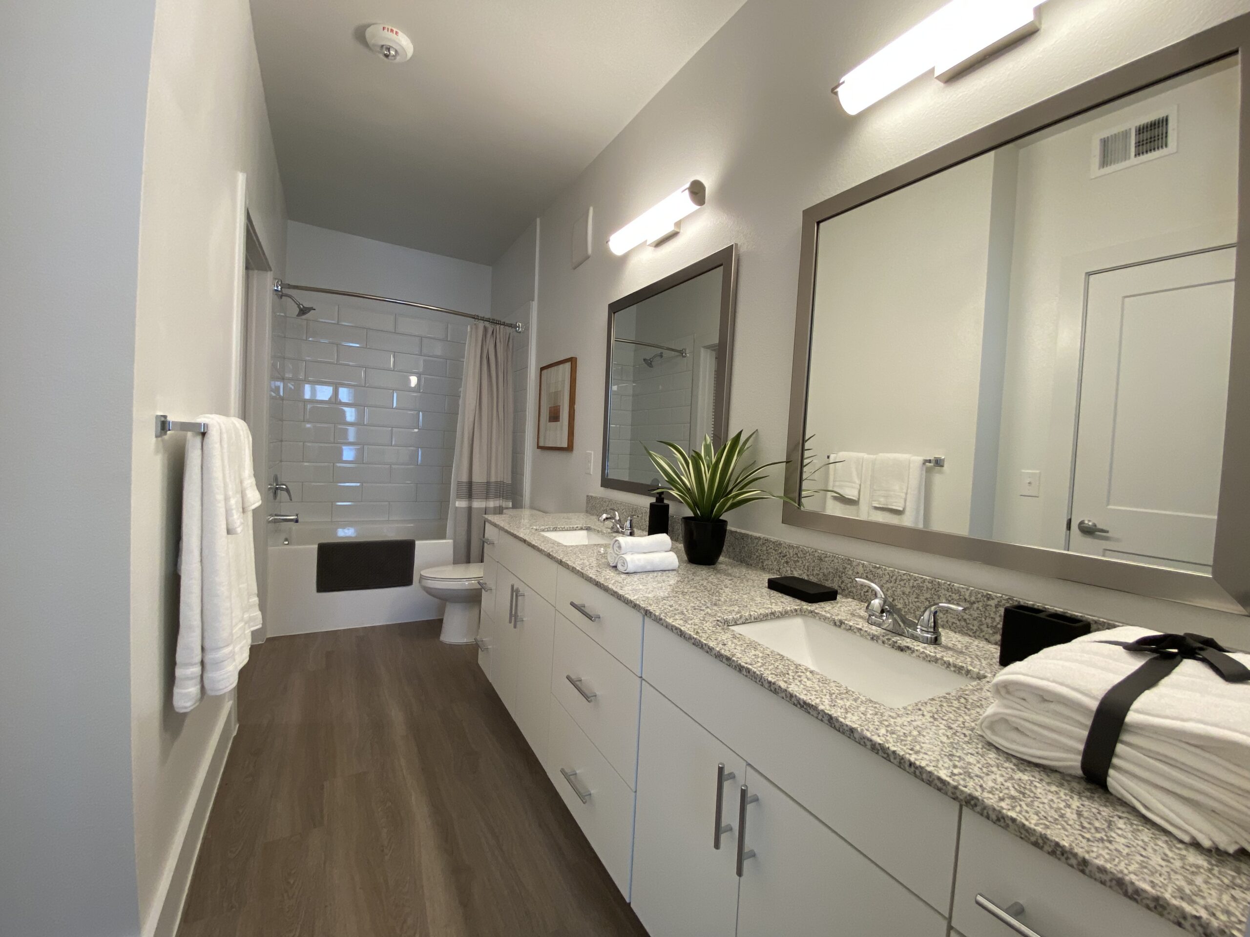 A modern bathroom features a double vanity with a granite countertop, two sinks, and stylish faucets. Above the vanity are two rectangular mirrors with sleek frames. A potted plant adds a touch of greenery. White towels are neatly arranged, and a few rolled towels are placed beside a black container. The bathroom includes a shower with a glass door and a bathtub, both surrounded by light gray subway tiles. The flooring is a warm wood tone, and the walls are painted in a soft gray.