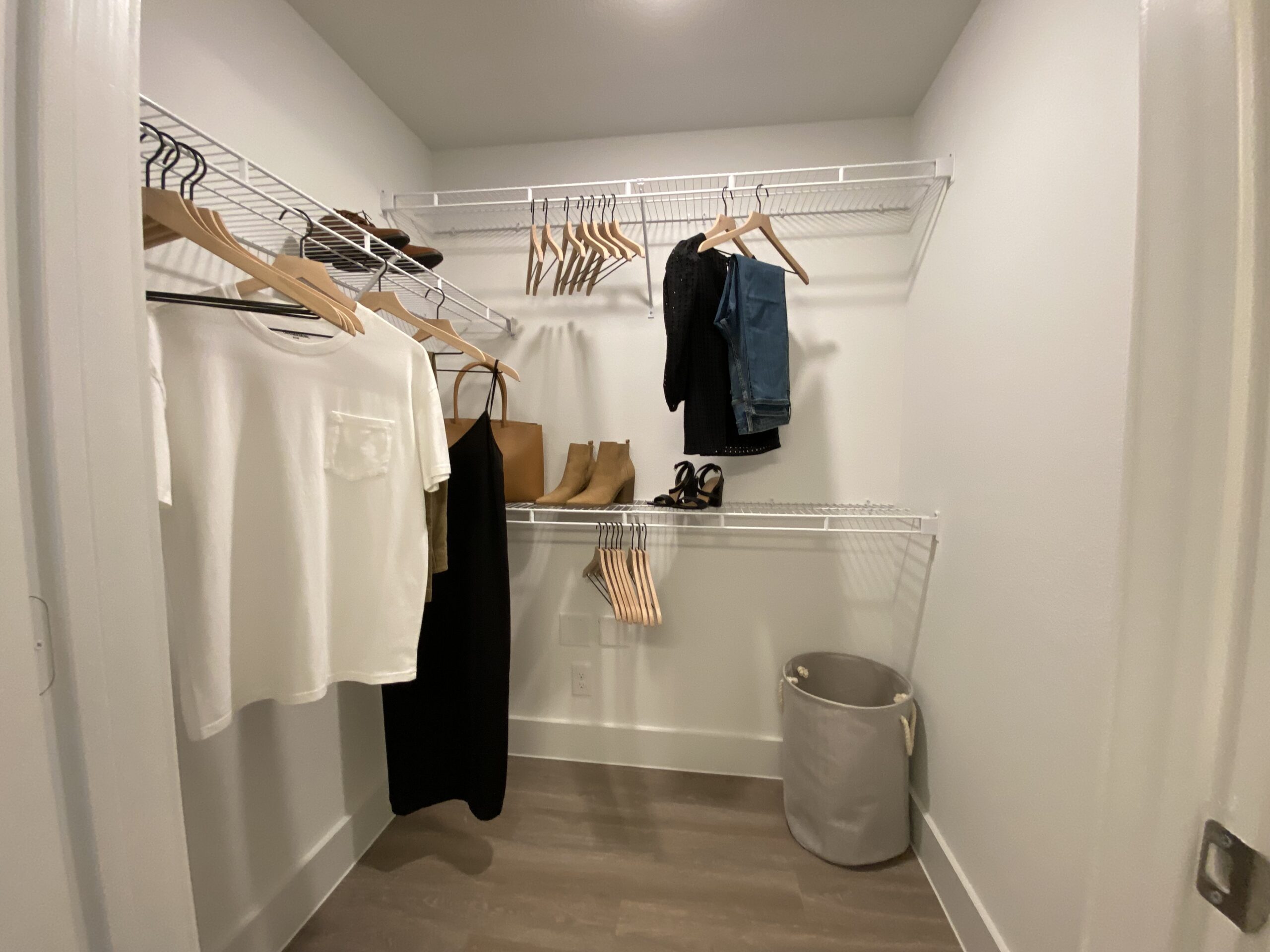 A well-organized closet features several clothing items hanging on wire shelves. On the left, a white t-shirt and a green top are visible, along with a long black dress hanging below. To the right, a pair of blue jeans and a black top are displayed. Footwear includes tan ankle boots and black high-heeled sandals on the floor. A brown tote bag is also hanging, and a gray laundry basket is positioned in the corner. The closet has a clean, minimalist aesthetic with light-colored walls and wooden flooring.