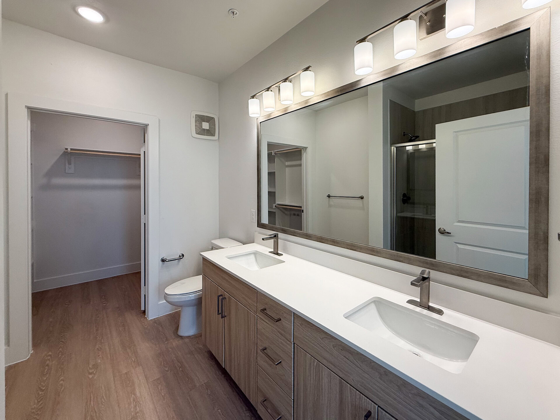 A modern San Antonio apartment bathroom features a double vanity with a white countertop and two rectangular sinks. A standing glass shower is in the background, and the flooring is a warm wood tone, enhancing the contemporary aesthetic.