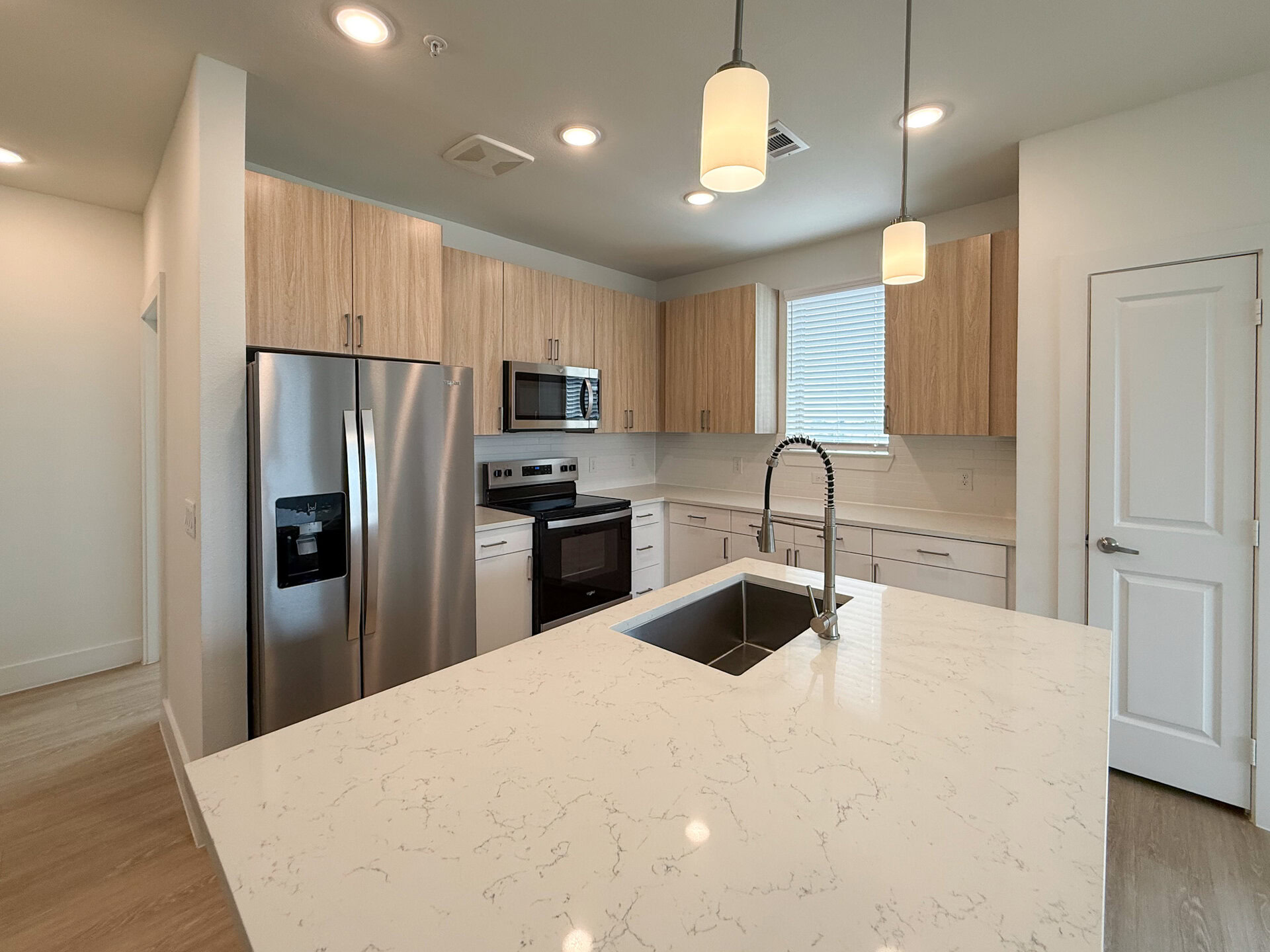 A modern San Antonio apartment kitchen featuring light wood dual-tone cabinetry and stainless steel appliances. A large island with a white marble countertop includes a deep sink with a sleek, pull-down faucet. Natural light enters through a window with blinds. The flooring is light-colored wood, contributing to the contemporary aesthetic.