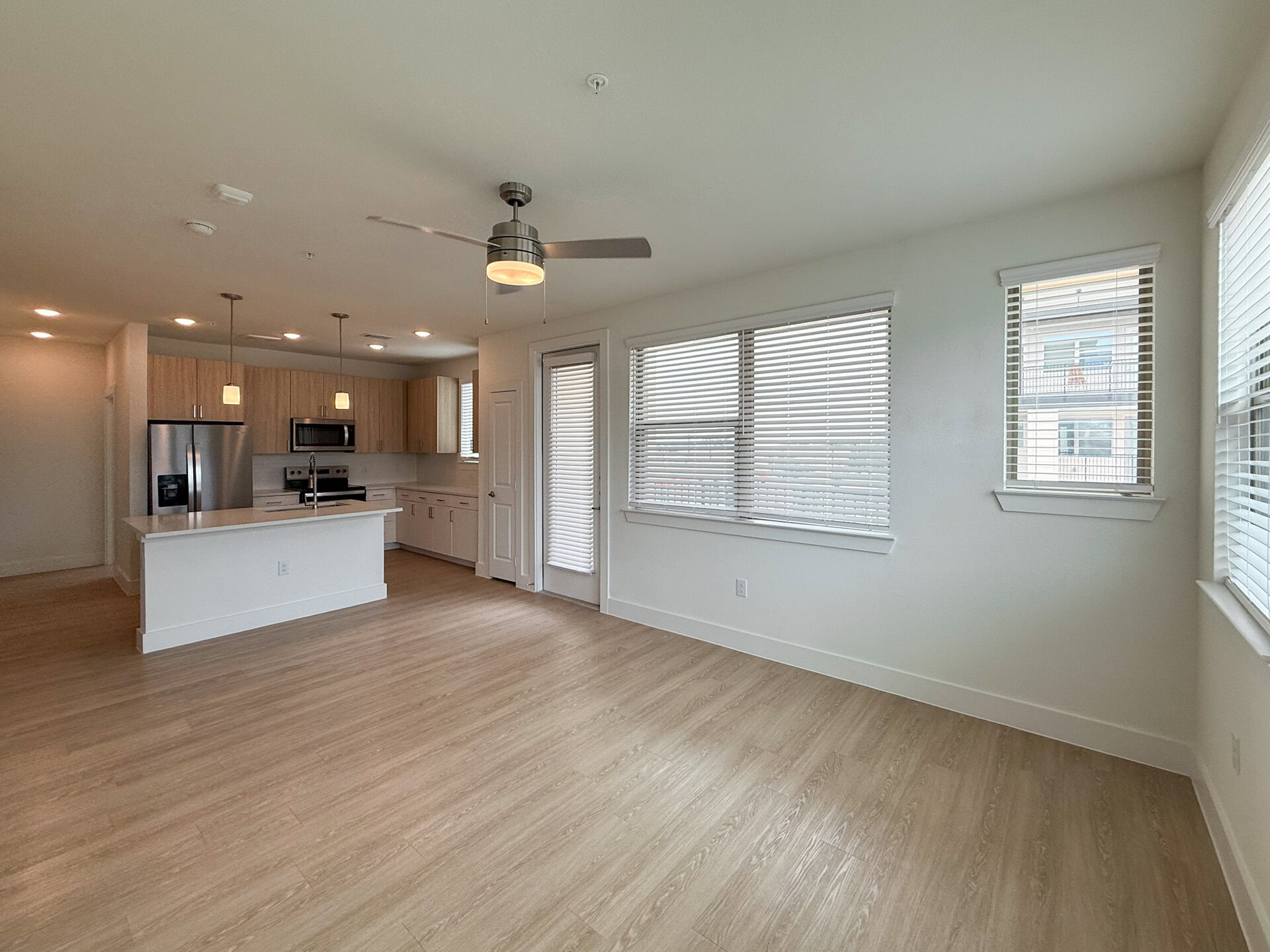 A modern, open-concept living room features a kitchen with light wood cabinetry, stainless steel appliances, and a central island with a white countertop. Pendant lights hang above the island. The living area has large windows with white blinds, allowing natural light to fill the room. The flooring is light-colored, resembling wood, and the walls are painted white, creating a bright and airy atmosphere.