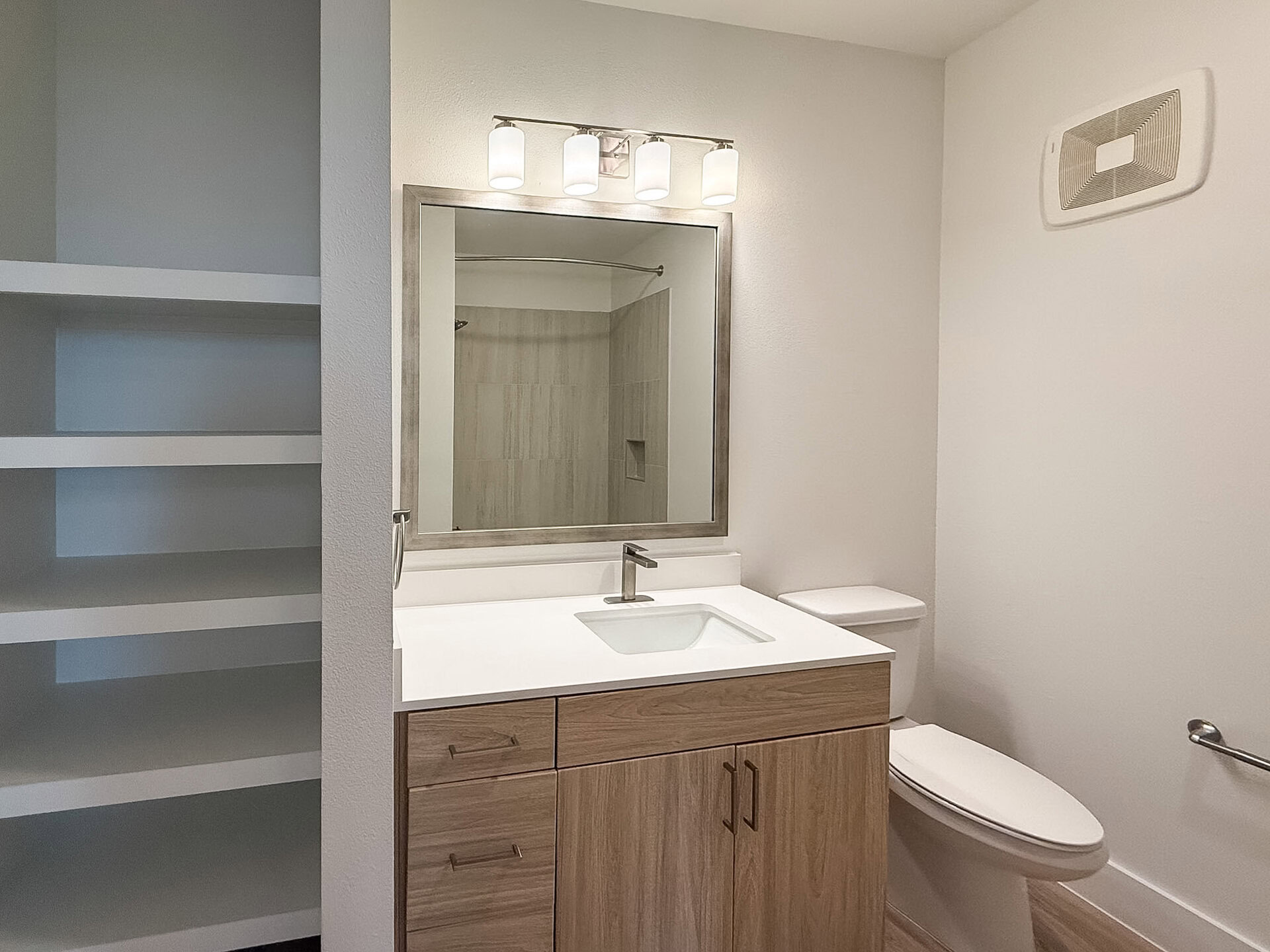 A modern San Antonio apartment bathroom features a double vanity with a white countertop and two rectangular sinks. A standing glass shower is in the background, and the flooring is a warm wood tone, enhancing the contemporary aesthetic.