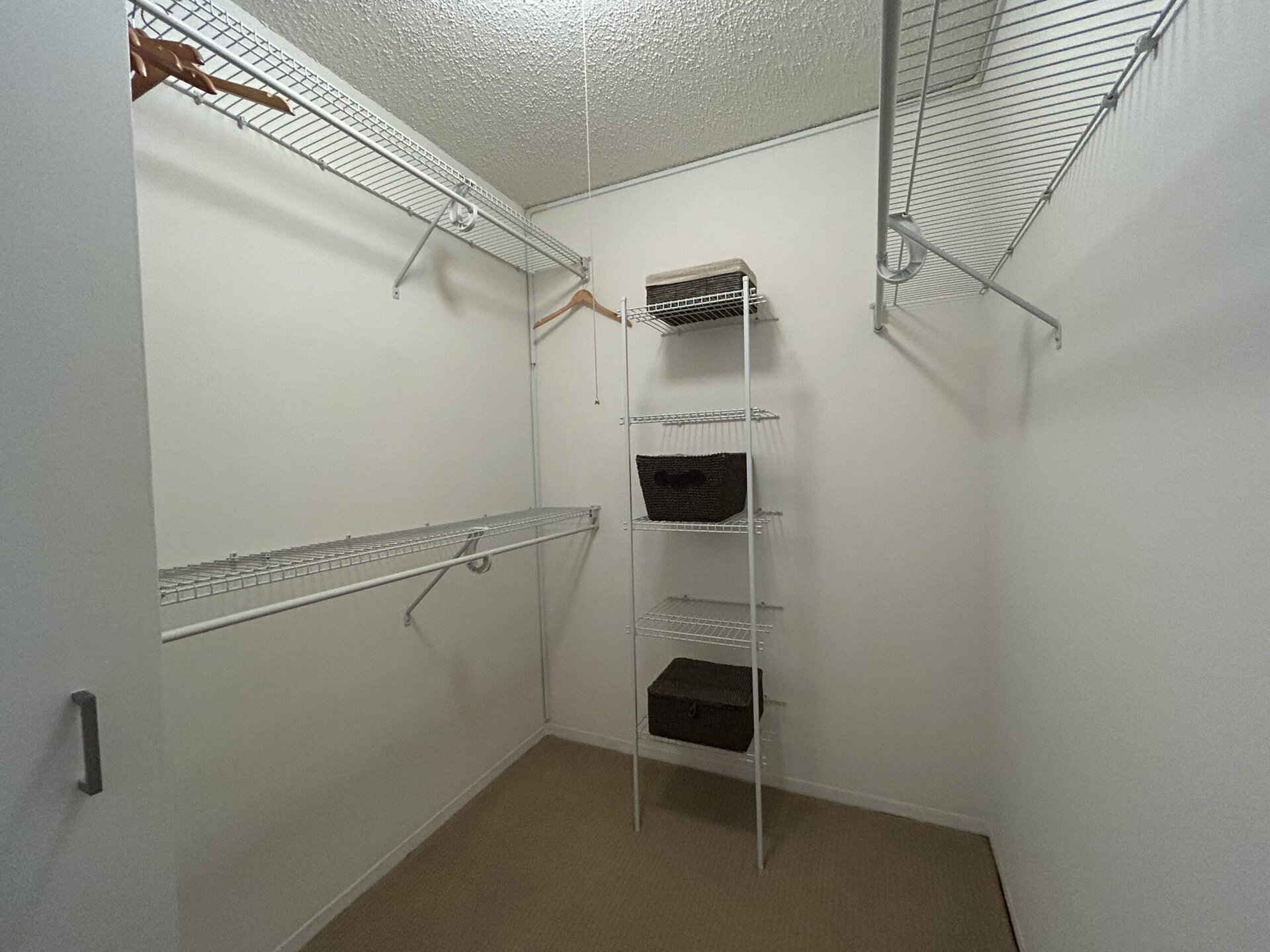 A spacious closet features white wire shelving and a hanging rod. On one side, there is a single hanger attached to the wall. Opposite, a tall shelving unit holds three woven baskets, with one basket on the top shelf and two below it. The walls are painted white, and the floor is carpeted in a neutral tone. The overall appearance is organized and minimalistic.