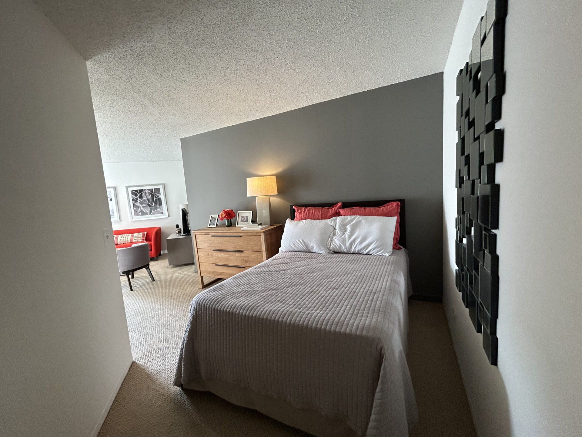 A cozy bedroom features a bed with a light gray quilt and white pillows, complemented by red accent pillows. Beside the bed is a wooden dresser with a lamp and decorative items, including a small vase of flowers and framed photos. The wall behind the bed is painted dark gray, while a textured black wall art piece adds visual interest. In the background, a living area is visible with a red sofa and a gray chair, creating a warm and inviting atmosphere. The flooring is carpeted, enhancing the room's comfort.
