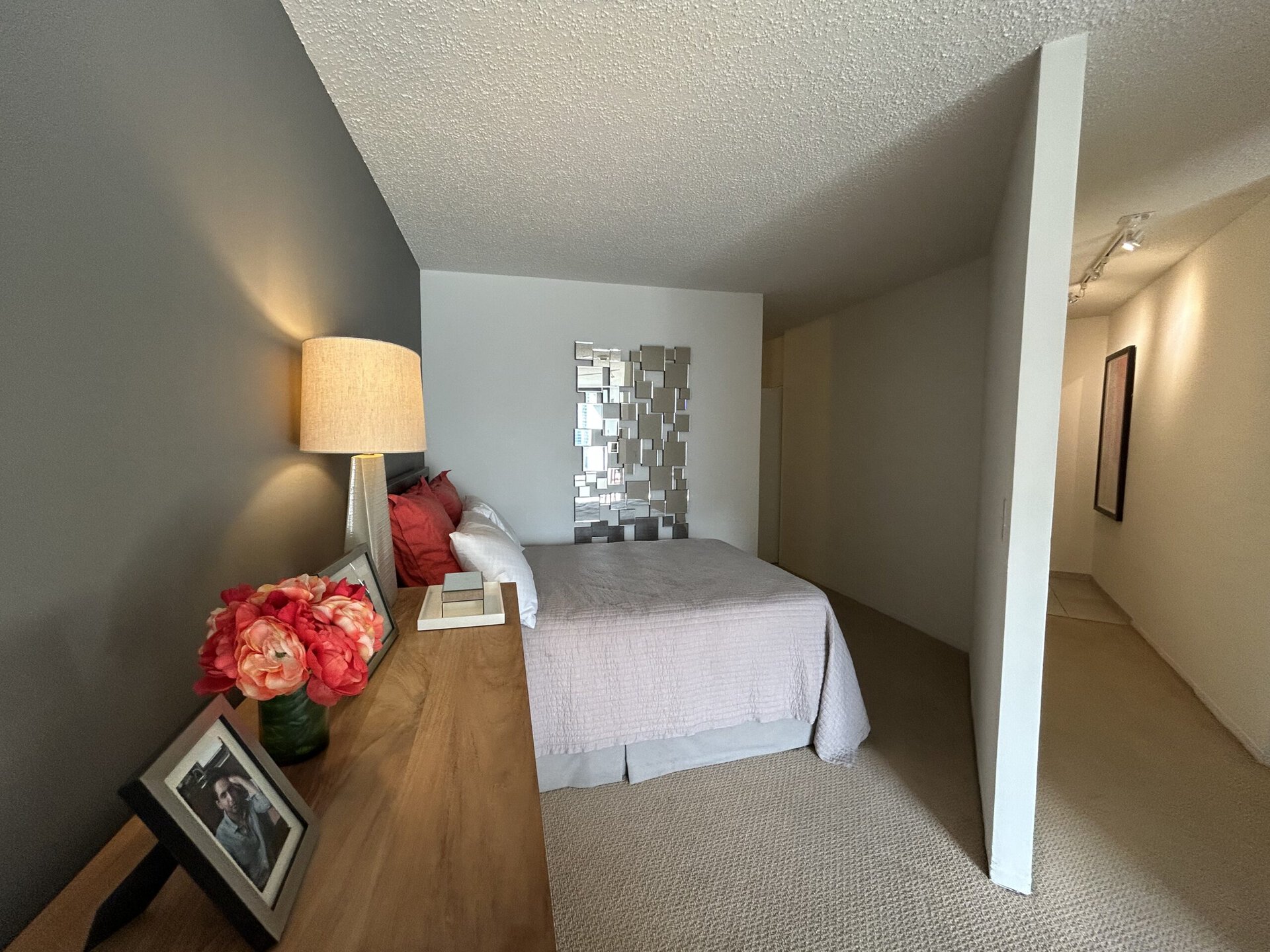 A cozy bedroom features a bed with a light pink quilt and white pillows, positioned against a dark gray wall. A decorative mirror made of small, reflective squares hangs above the bed. To the left, a wooden dresser holds a vase of pink flowers and framed photographs. Soft lighting from a lamp on the dresser creates a warm ambiance. The room has a neutral carpet and leads to a hallway with soft lighting and a framed artwork on the wall.