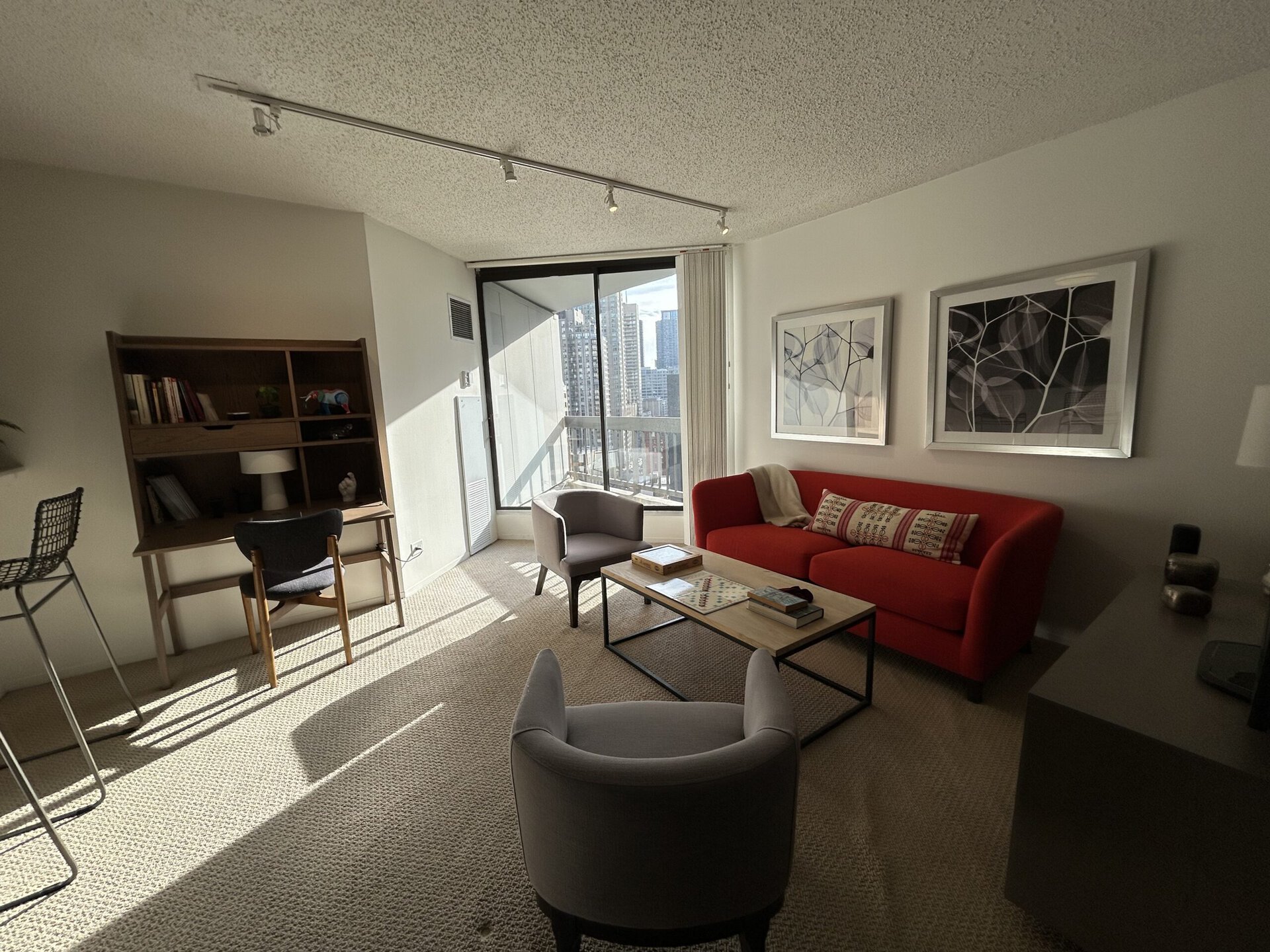 A modern living room features a bright, red sofa adorned with patterned cushions, complemented by a light gray armchair. A wooden coffee table sits in the center, displaying a few books and a board game. Behind the sofa, two framed artworks hang on the wall. A wooden bookshelf with a lamp and decorative items is positioned nearby, alongside a tall, metal barstool. Large windows allow natural light to fill the space, showcasing a cityscape view outside. The room has a cozy, inviting atmosphere with neutral-toned carpeting.