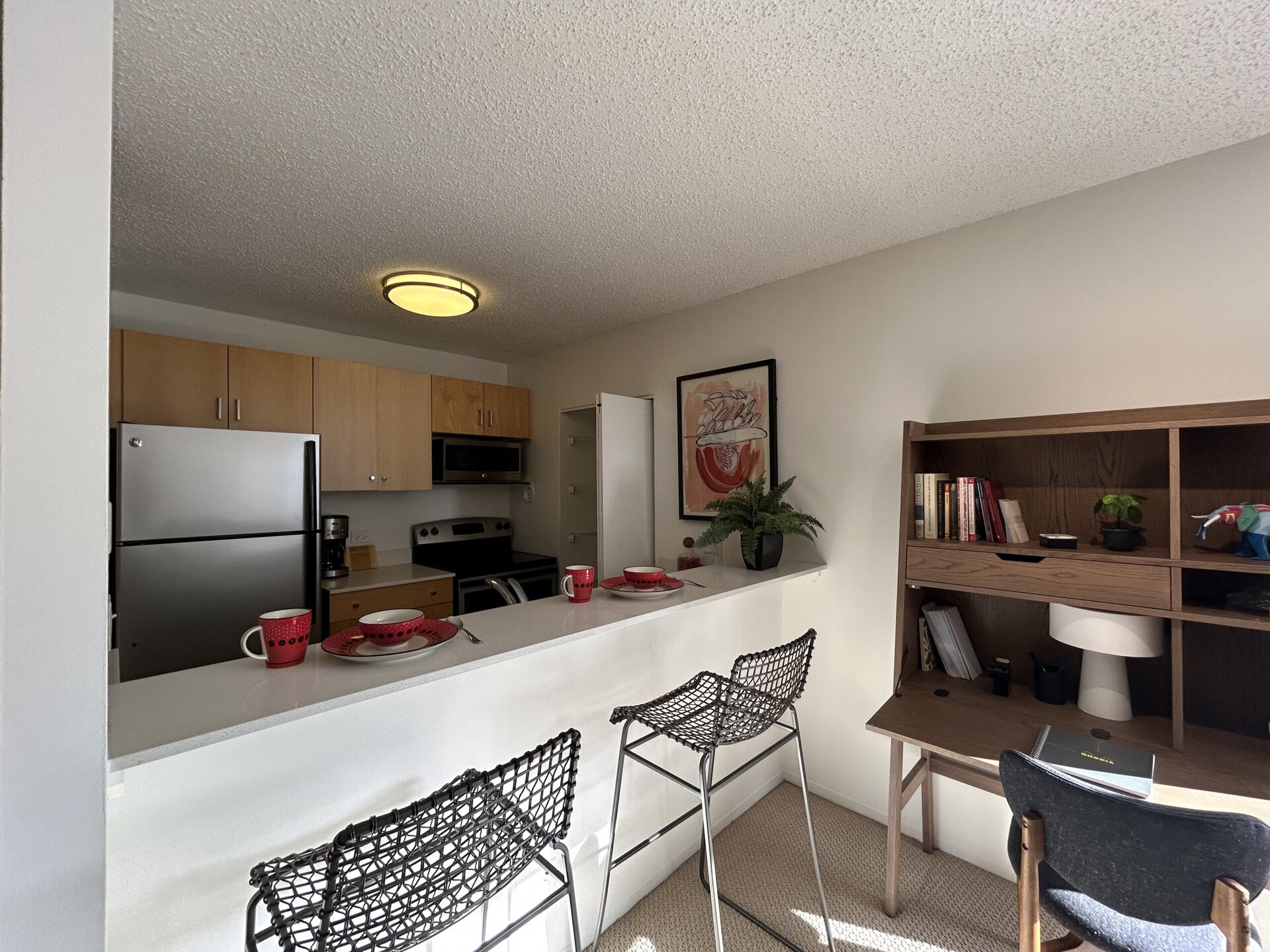A modern kitchen area features light wood cabinetry and stainless steel appliances, including a refrigerator and an oven. A white countertop serves as a breakfast bar with two black wire stools. On the counter, there are two red mugs and a bowl on a decorative plate. A potted fern adds a touch of greenery. In the background, a wooden desk holds books, a lamp, and a small plant, creating a cozy workspace. A piece of artwork hangs on the wall, adding color to the space.