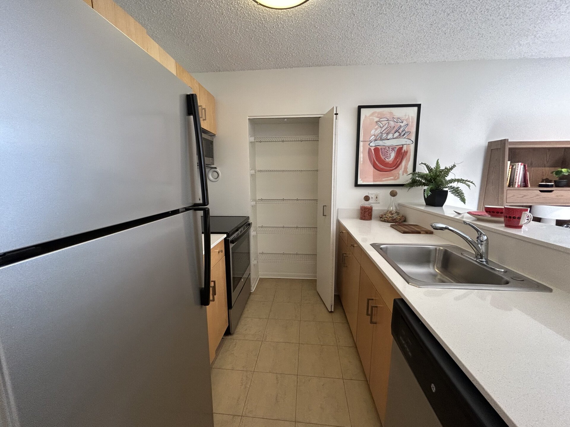 A modern kitchen features a stainless steel refrigerator and an oven with a black cooktop. The countertop is white and includes a stainless steel sink. An open pantry with wire shelves is visible, and a framed artwork hangs on the wall. A small potted fern adds greenery, while two red mugs and a wooden cutting board are placed on the counter. The cabinetry is light wood, creating a bright and inviting atmosphere.