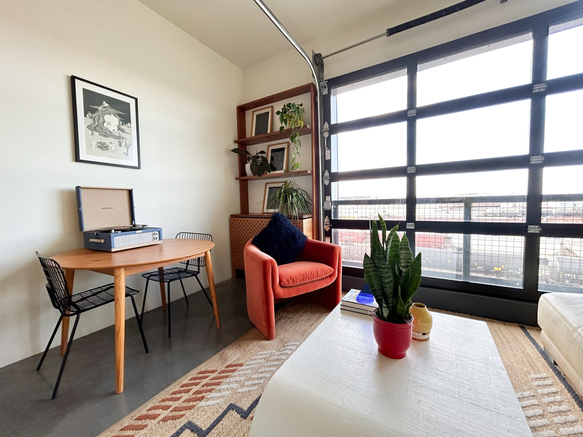 A cozy corner of a modern living space features a round wooden table with two black wire chairs. On the table sits a vintage-style record player with its lid open. Nearby, a plush orange armchair with a dark blue cushion invites relaxation. A shelf above the table displays framed artwork and potted plants, adding greenery to the room. A large glass garage door allows natural light to flood in, revealing a view of the outside. A potted snake plant and a small decorative vase are placed on a light-colored rug in front of the armchair.