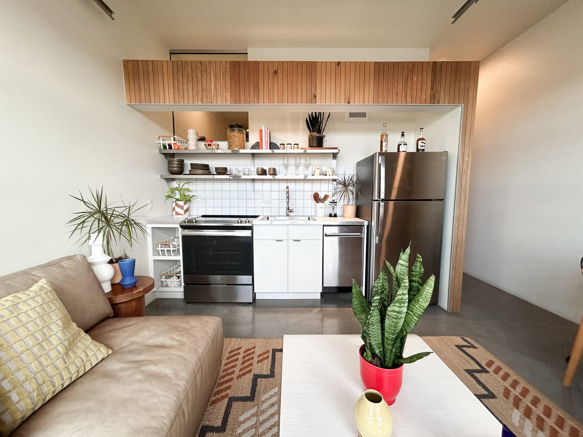 A modern kitchen area features a combination of stainless steel and white appliances, including a refrigerator, oven, and dishwasher. Above the countertop, open shelving displays various dishes, glasses, and decorative items. A small potted plant sits on the counter, alongside utensils and a sink. The kitchen is partially separated from a cozy living area with a light brown sofa adorned with a patterned cushion. A coffee table with a red pot containing a tall plant is positioned in front of the sofa, and a textured rug adds warmth to the space.