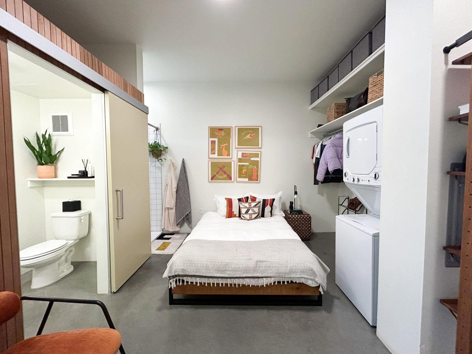 A compact living space features a bed with decorative pillows and a light gray throw blanket. Above the bed, there are four framed art pieces on the wall. To the left, a small bathroom area is partially visible, showcasing a toilet and a potted plant. Adjacent to the bed, a stacked washer and dryer unit is present, along with a shelf holding clothing and storage baskets. The floor is concrete, and there are hanging towels and a small rug near the bathroom entrance.
