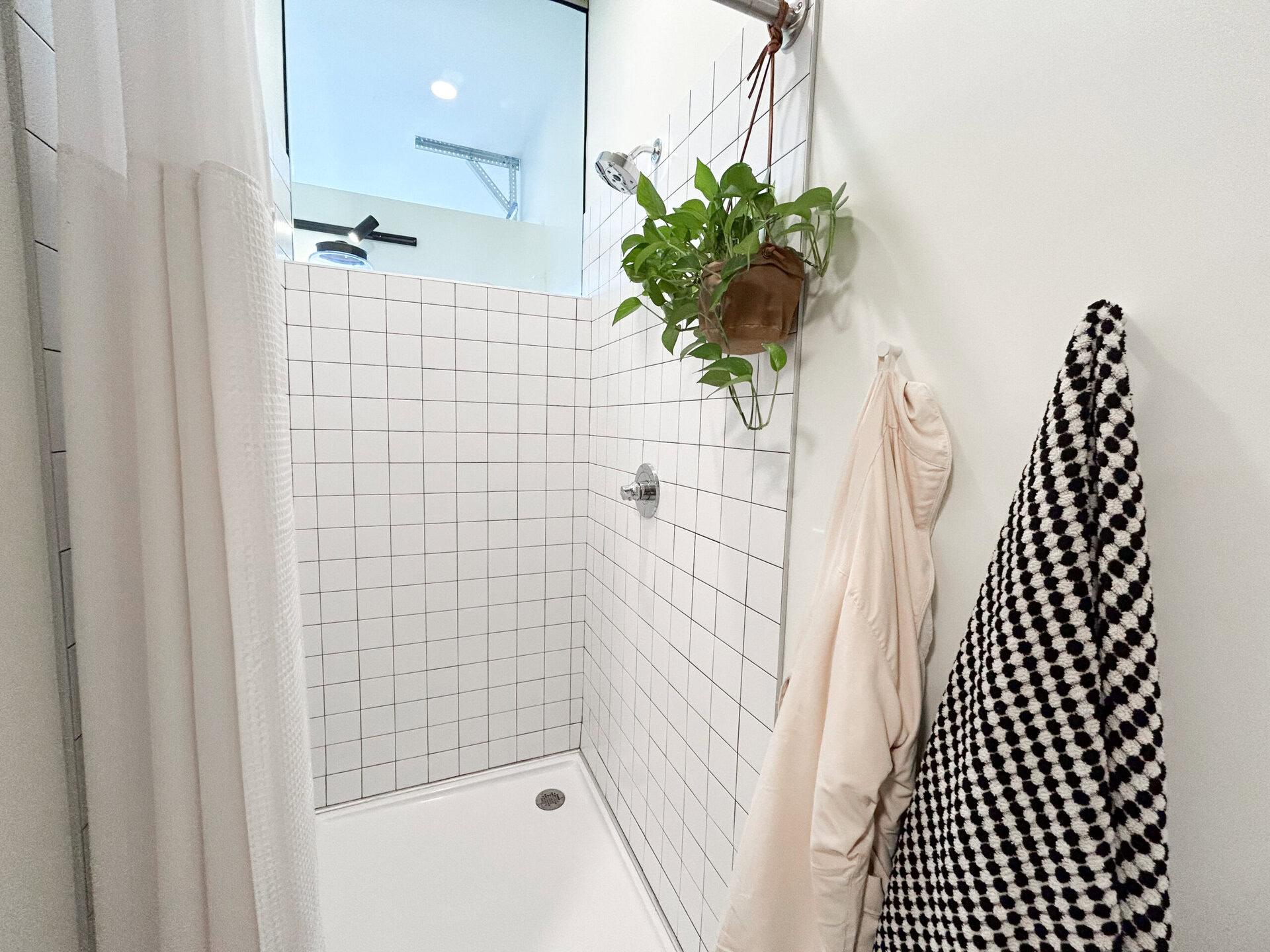 A modern shower space features white tiled walls with a grid pattern. A chrome showerhead and control handle are mounted on one wall. A potted plant hangs from the ceiling, adding a touch of greenery. A white shower curtain is partially visible, and two towels are hanging on hooks: one is a light color, while the other has a black and white polka dot pattern. The overall aesthetic is clean and contemporary.