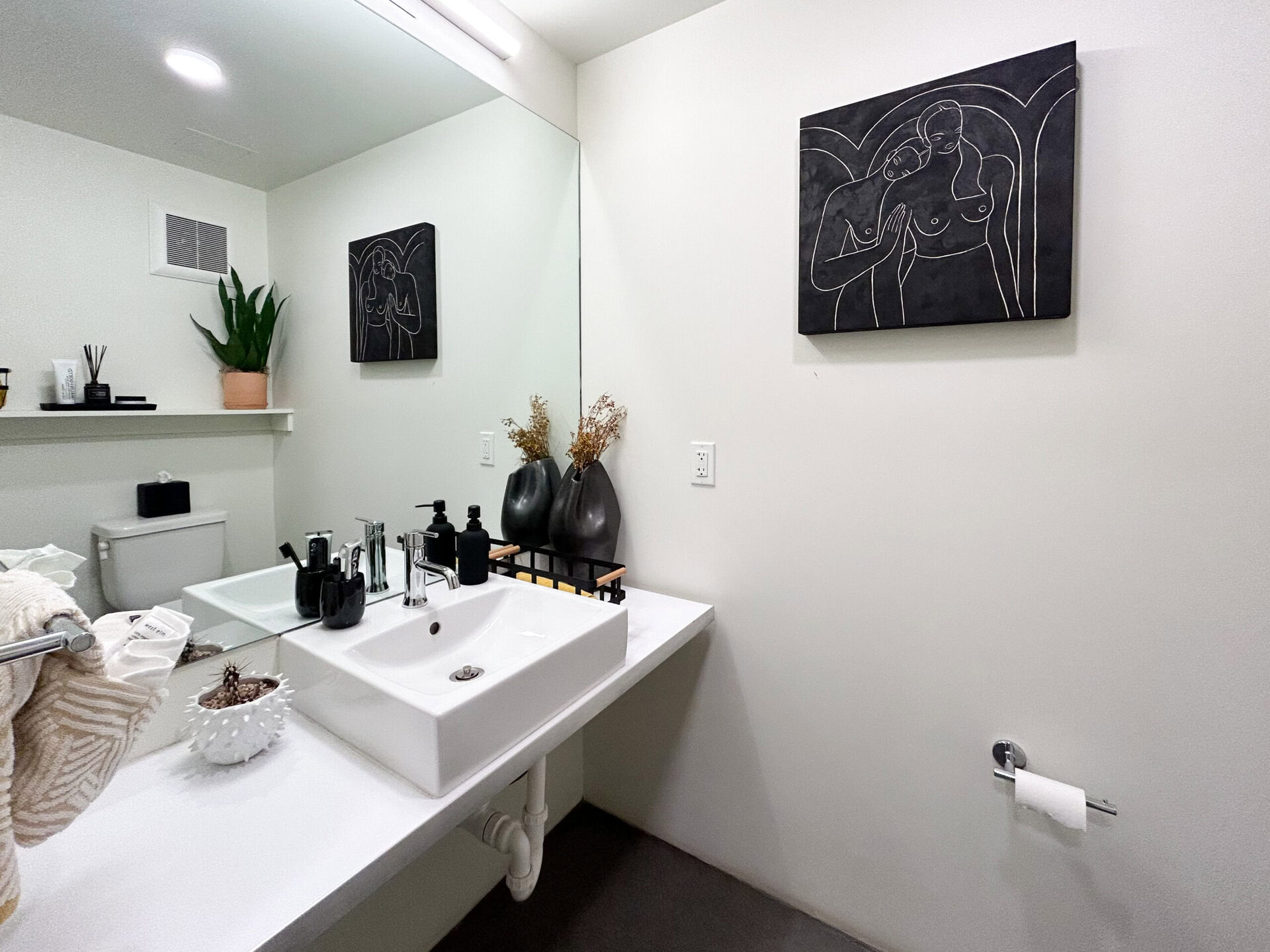 A modern bathroom features a sleek white sink with a chrome faucet, positioned on a white countertop. Above the sink, a large mirror reflects the space. To the left, a toilet is visible, while a small shelf holds decorative items, including a black container and a potted plant. On the right wall, a black artwork depicting abstract figures is displayed. Two dark vases with dried flowers sit on the countertop alongside various black bottles and a small decorative bowl. A towel hangs on a chrome rack near the sink.