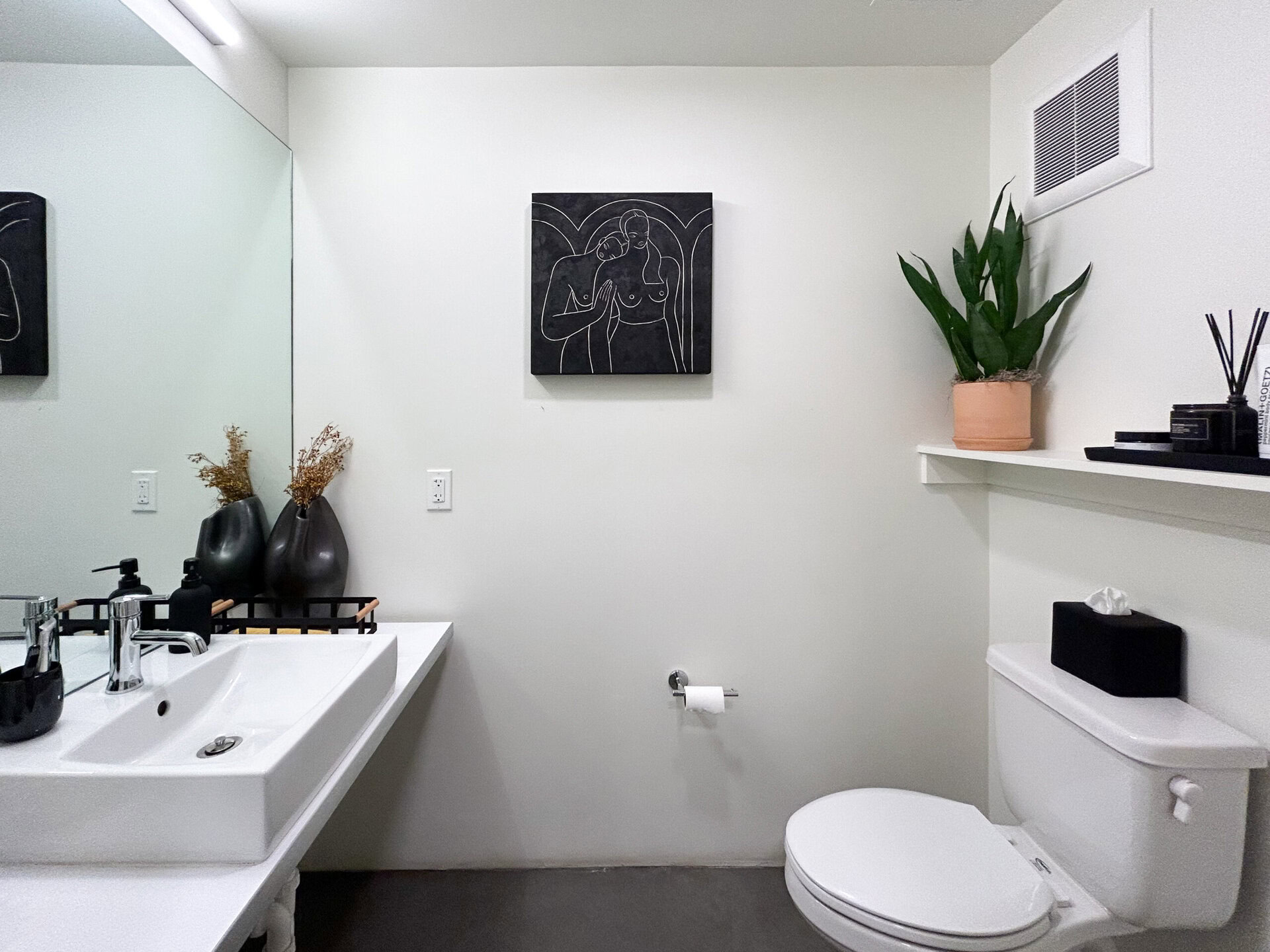 A modern bathroom features a sleek white sink with a chrome faucet and a black soap dispenser. Above the sink, a large mirror reflects the space. On the wall, there is a black artwork depicting two figures outlined in white. To the right, a shelf holds a potted plant, a black candle, and a tissue box. Two black vases with dried flowers are placed on a wooden tray beside the sink. A toilet with a roll of toilet paper is positioned in the corner. The walls are painted a soft white, creating a clean and minimalist aesthetic.