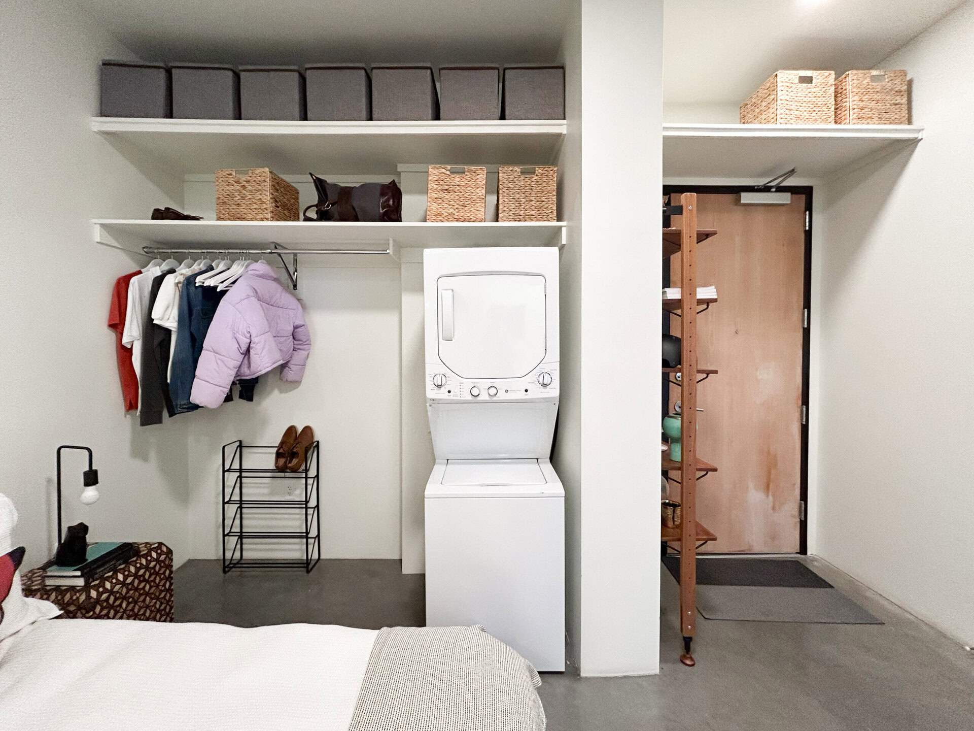 A compact laundry area features a stacked washer and dryer unit against a wall. To the left, a clothing rack holds several garments, including a purple puffer jacket and various shirts. Above the rack, there are gray storage boxes and woven baskets for organization. A black metal shoe rack sits below the clothing, displaying a pair of brown shoes. In the foreground, a bedside table with a patterned design holds a stack of books and a small lamp. The space is well-lit and has a minimalist aesthetic, with a wooden door visible in the background.