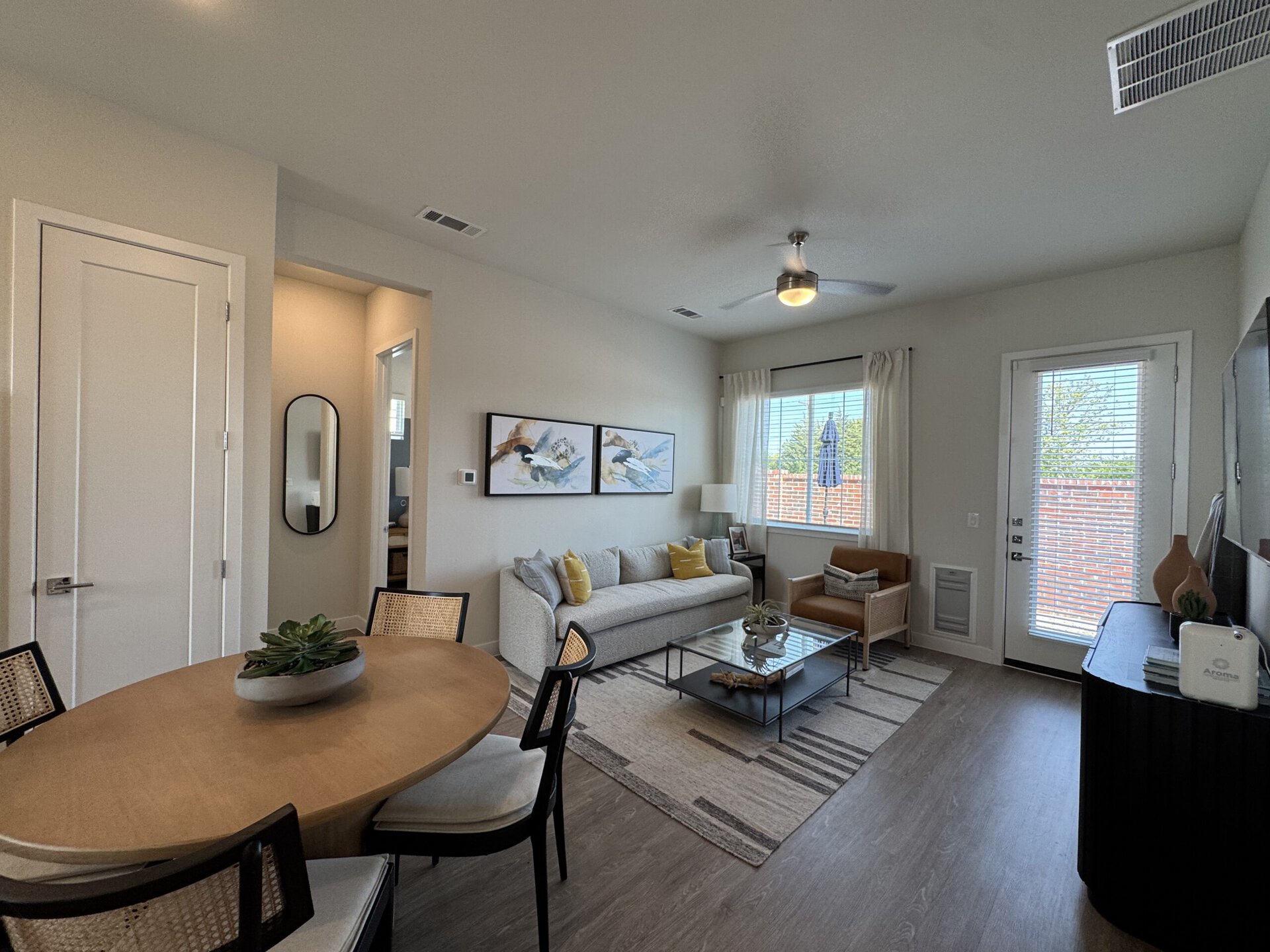 A modern living space featuring a light-colored sofa adorned with decorative pillows, positioned next to a glass coffee table. Two framed artworks hang on the wall above the sofa. A cozy armchair in a warm tone complements the seating area. A round dining table with black chairs is visible, topped with a decorative plant. Natural light streams in through a window with blinds, revealing a view of greenery outside. The room has a neutral color palette, with a ceiling fan and light fixtures adding to the ambiance.
