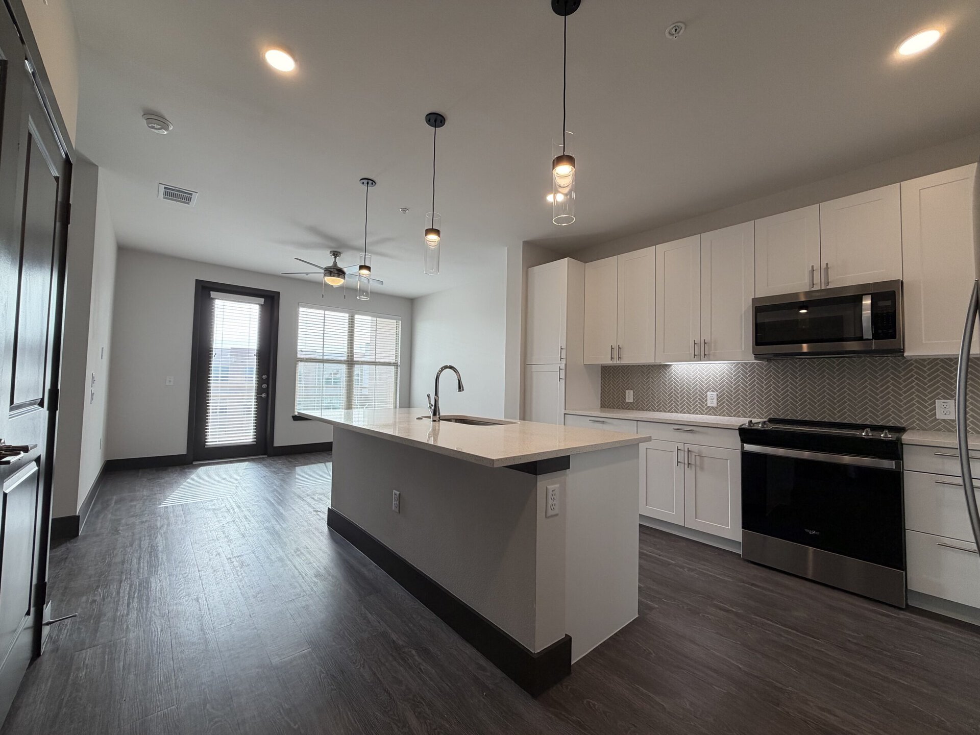 A modern kitchen space features a large island with a light-colored countertop and a sink. Above, pendant lights hang from the ceiling. The cabinetry is sleek and white, complemented by a herringbone-patterned backsplash. A stainless steel microwave and oven are integrated into the cabinetry. To the side, a door leads to a balcony, with large windows allowing natural light to fill the room. The flooring is dark and wood-like, enhancing the contemporary aesthetic.