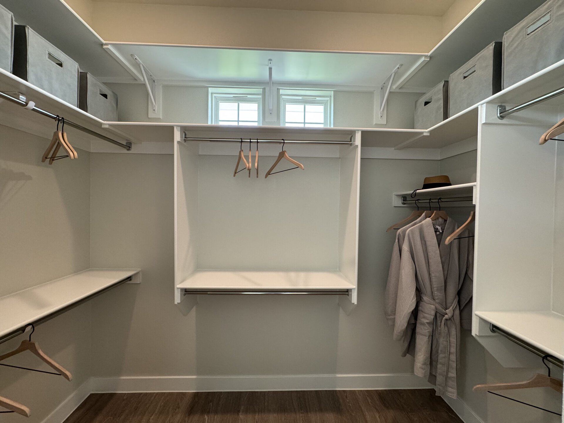 A spacious, organized closet features white shelving and a hanging rod. Several wooden hangers are visible, with two gray robes hanging on the right side. A brown hat rests on a shelf above the robes. Gray storage boxes are neatly arranged on the upper shelves. Natural light filters in through two small windows at the top. The overall design is clean and minimalist.