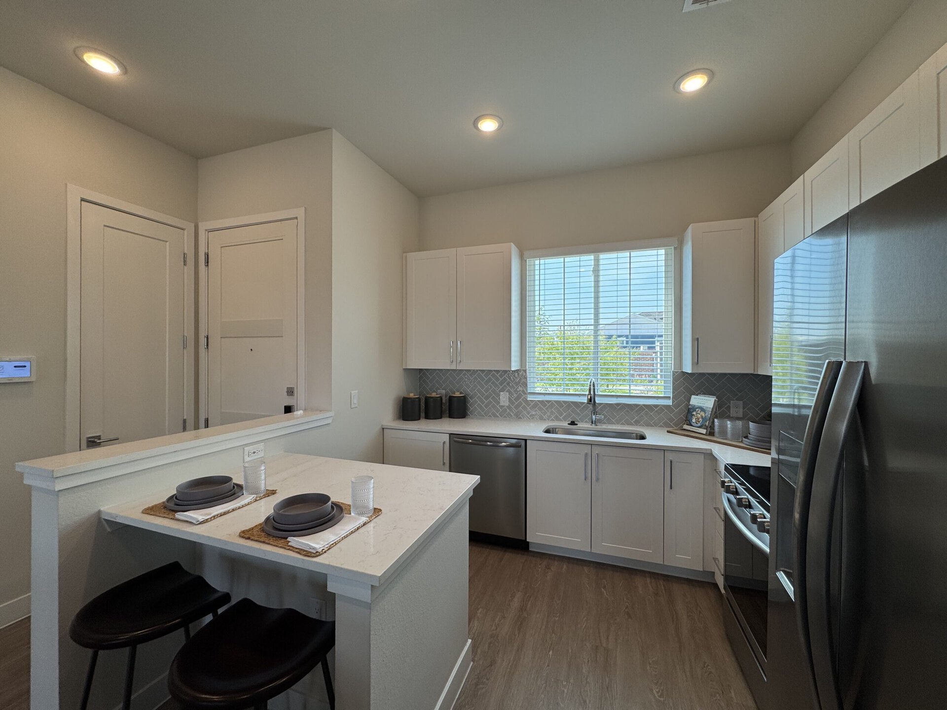 A modern kitchen features light-colored cabinetry and a sleek countertop. The kitchen includes stainless steel appliances, including a refrigerator and a dishwasher. A window above the sink allows natural light to enter, with blinds partially drawn. On the countertop, there are two dark plates on woven placemats, accompanied by white napkins and a glass. The backsplash is designed with a herringbone pattern, and there are decorative black containers on the counter. The overall aesthetic is clean and contemporary.
