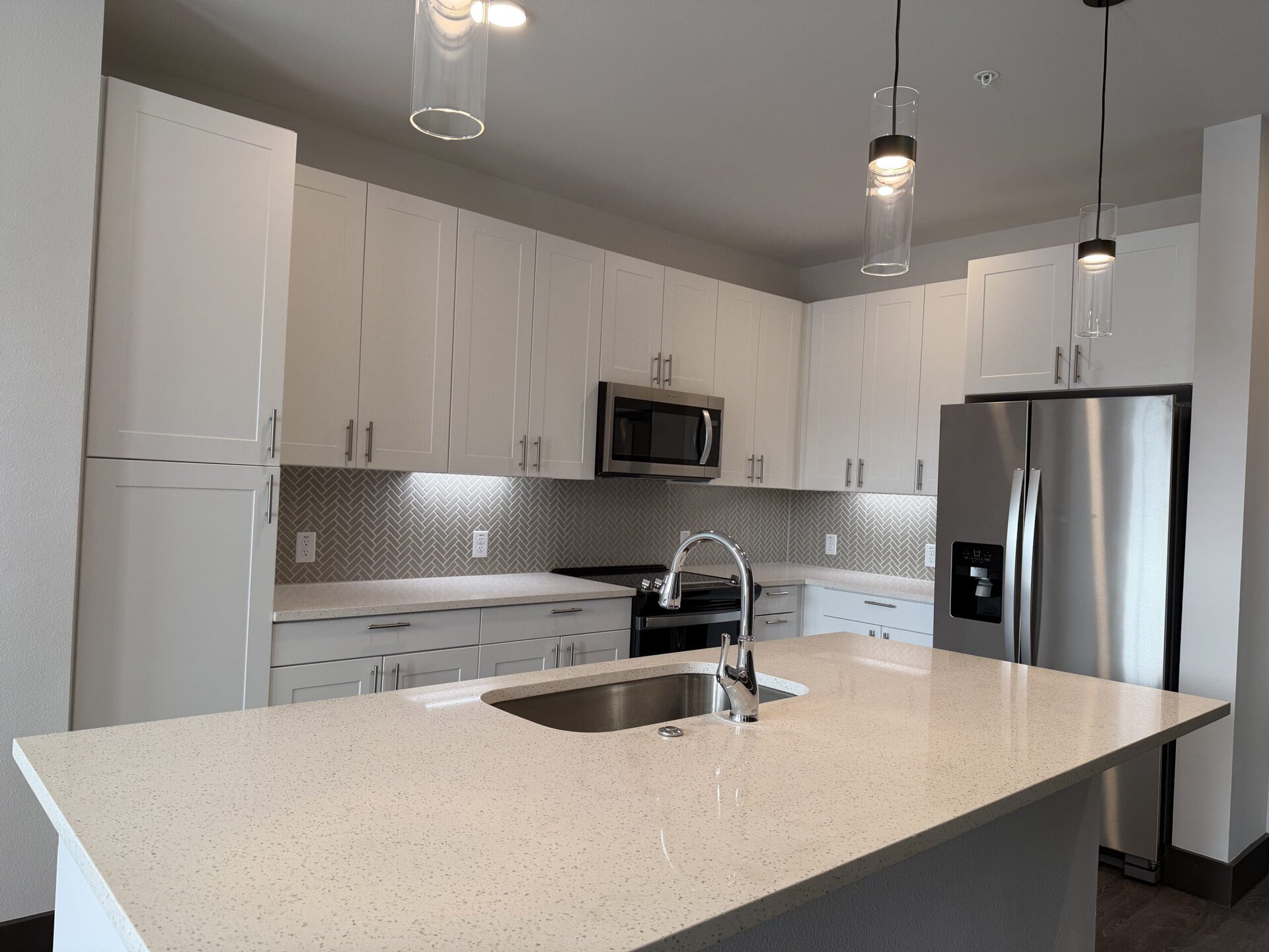 A modern kitchen featuring white cabinetry with sleek handles, a stainless steel microwave, and a black stove. The backsplash has a herringbone pattern in light tones. A large island with a white speckled countertop includes a stainless steel sink and a contemporary faucet. Two pendant lights hang above the island, providing illumination. A stainless steel refrigerator is positioned to the right. The overall design is bright and contemporary.