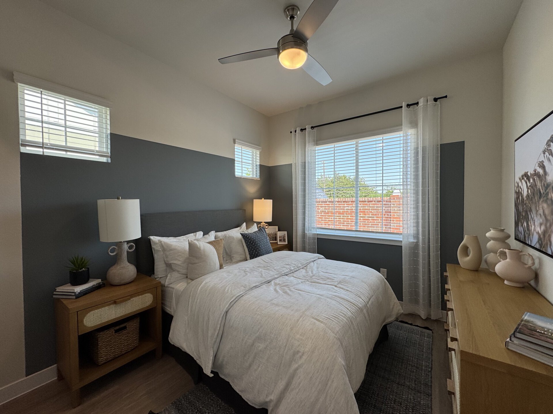 A cozy bedroom features a queen-sized bed with white bedding and multiple decorative pillows. The headboard is upholstered in gray fabric. A bedside table made of light wood holds a textured lamp and a small potted plant. Natural light streams in through a large window with white blinds, complemented by sheer white curtains. The walls are painted in a two-tone scheme, with the lower half in a dark gray. A wooden dresser with decorative vases and a framed picture is positioned against one wall, adding to the room's modern aesthetic.
