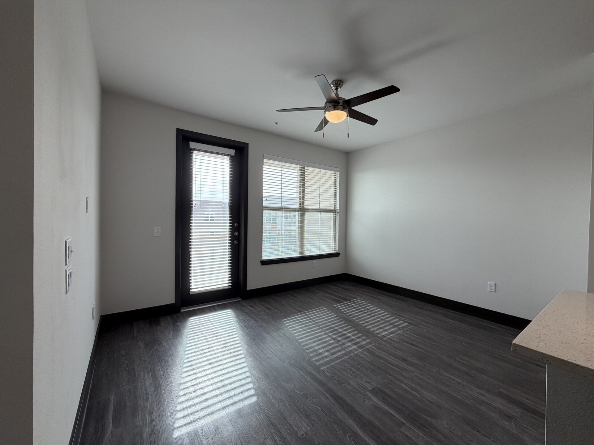 A spacious room features a ceiling fan with wooden blades and a light fixture. There are large windows with blinds allowing natural light to enter, casting shadows on the dark laminate flooring. A door leads to an outdoor area, and the walls are painted in a light color, creating a bright and airy atmosphere. A countertop is visible in the corner, suggesting a kitchen or bar area.
