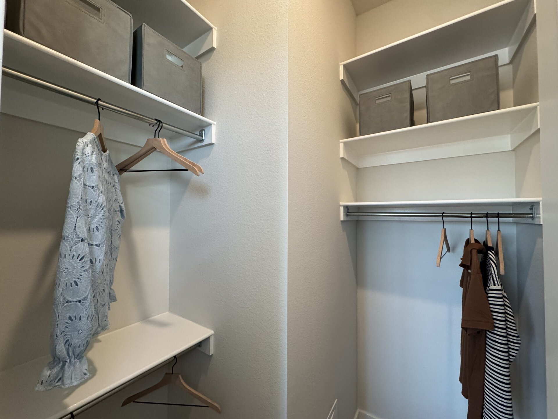 A well-organized closet features two sections. On the left, a light blue lace garment hangs from a wooden hanger, with an empty shelf below it. On the right, there are two gray storage boxes on a shelf above a hanging rod, which holds three garments: a brown shirt, a striped shirt, and another item. The walls are painted a light color, contributing to a clean and tidy appearance.