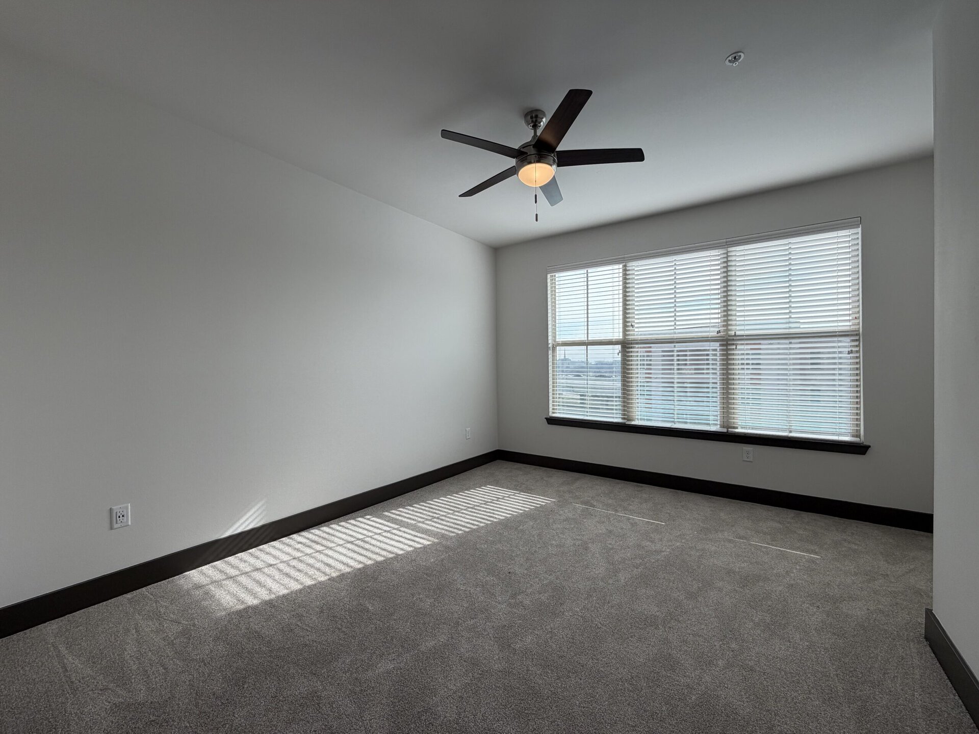 A spacious, empty room features light gray carpet and white walls. A ceiling fan with a light fixture hangs from the ceiling. Natural light streams in through a large window with horizontal blinds, casting shadows on the floor. The window is framed by a dark trim, contrasting with the lighter walls. The room has a clean, modern aesthetic, suitable for various uses.