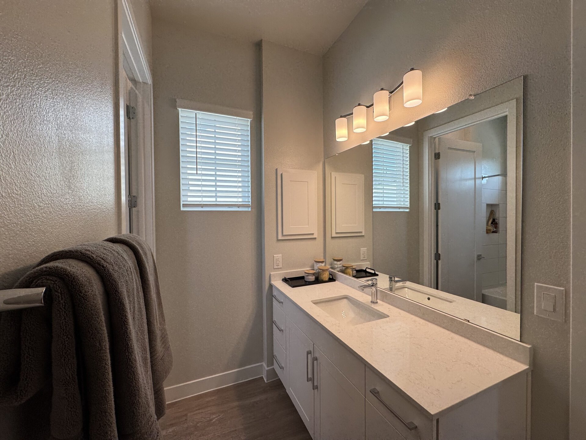 A modern bathroom features a sleek white vanity with a smooth countertop and an integrated sink. Above the vanity, three cylindrical light fixtures provide illumination. The wall is painted in a light neutral tone, and a large mirror reflects the space. To the left, a towel hangs on a chrome rack, while a window with horizontal blinds allows natural light to enter. A door leads to a shower area, partially visible in the background. The overall design is clean and contemporary.