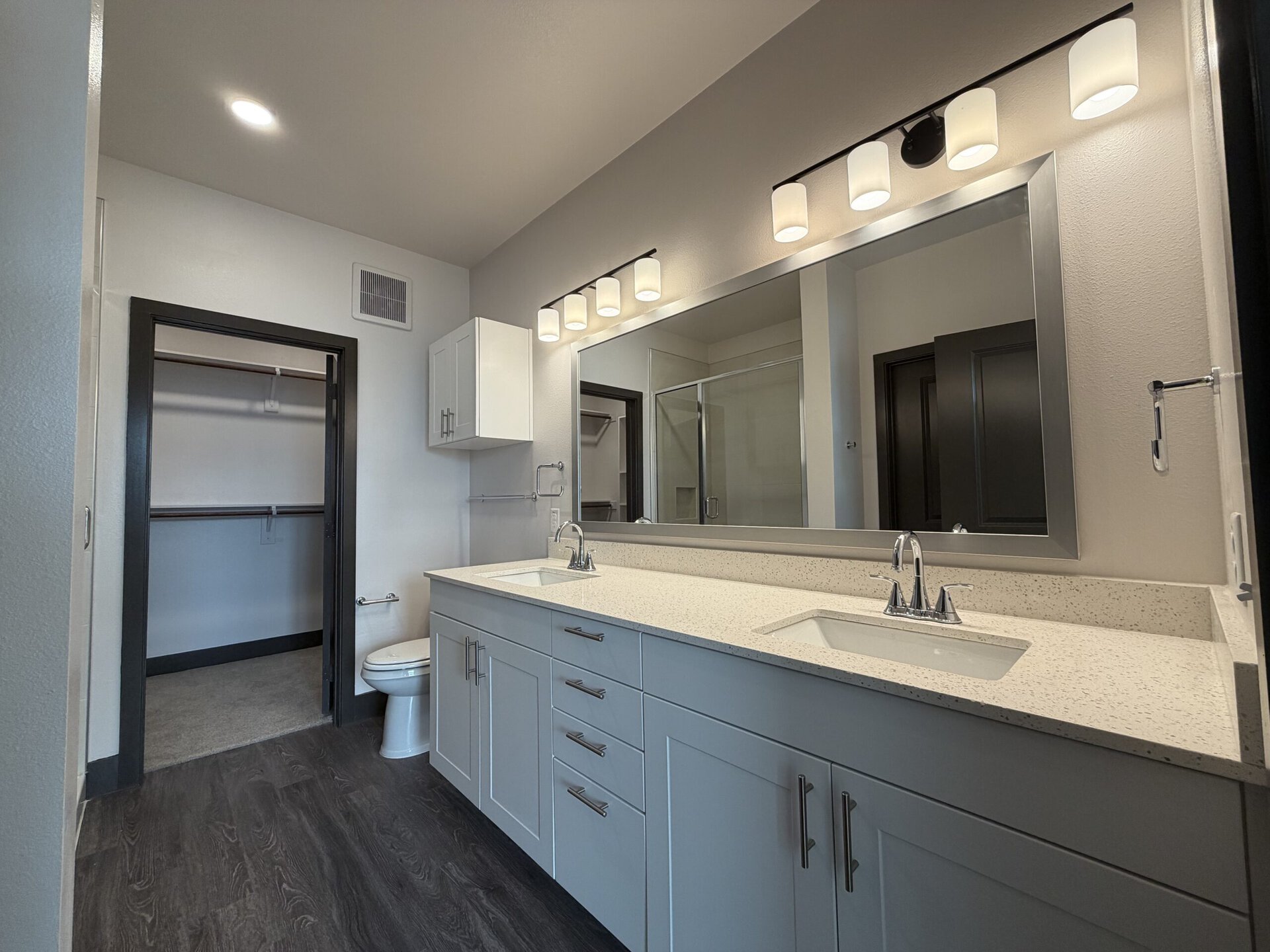 A modern bathroom features a double vanity with a light-colored speckled countertop and two sinks. Above the vanity, a large mirror is framed in silver, complemented by five cylindrical light fixtures. The cabinetry is sleek and white, with brushed metal handles. To the left, there is a closet with a sliding door, and a toilet is positioned nearby. The flooring is dark, contrasting with the light walls and cabinetry, creating a contemporary aesthetic. A glass shower enclosure is visible in the background.