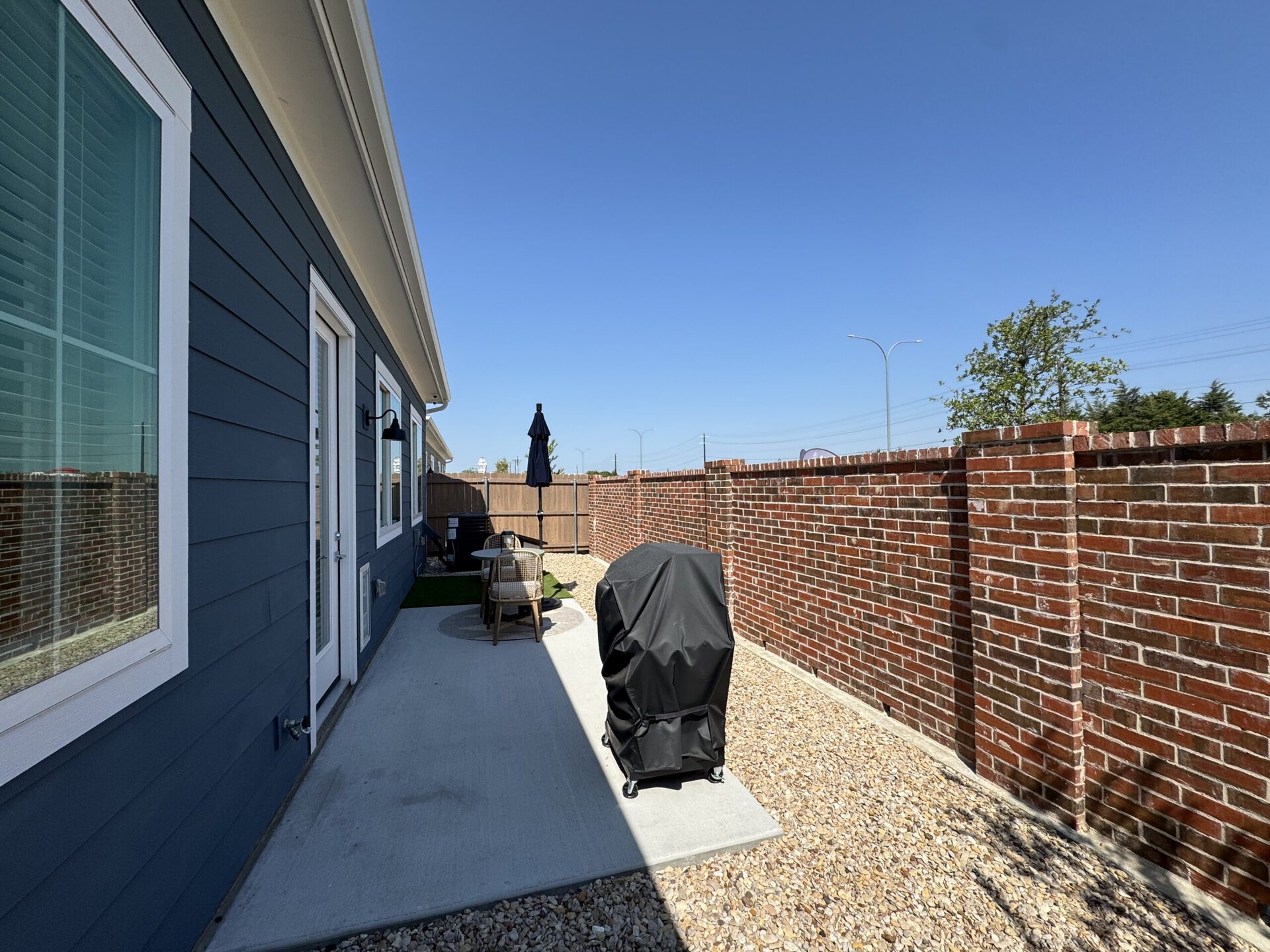 A narrow outdoor space features a blue house wall with large windows and a door. A black grill, covered with a protective cover, is positioned on a concrete path. Nearby, a small round table with two chairs sits under a black umbrella. The area is bordered by a tall brick wall, and there are patches of gravel on the ground. The sky is clear and blue, indicating a sunny day.