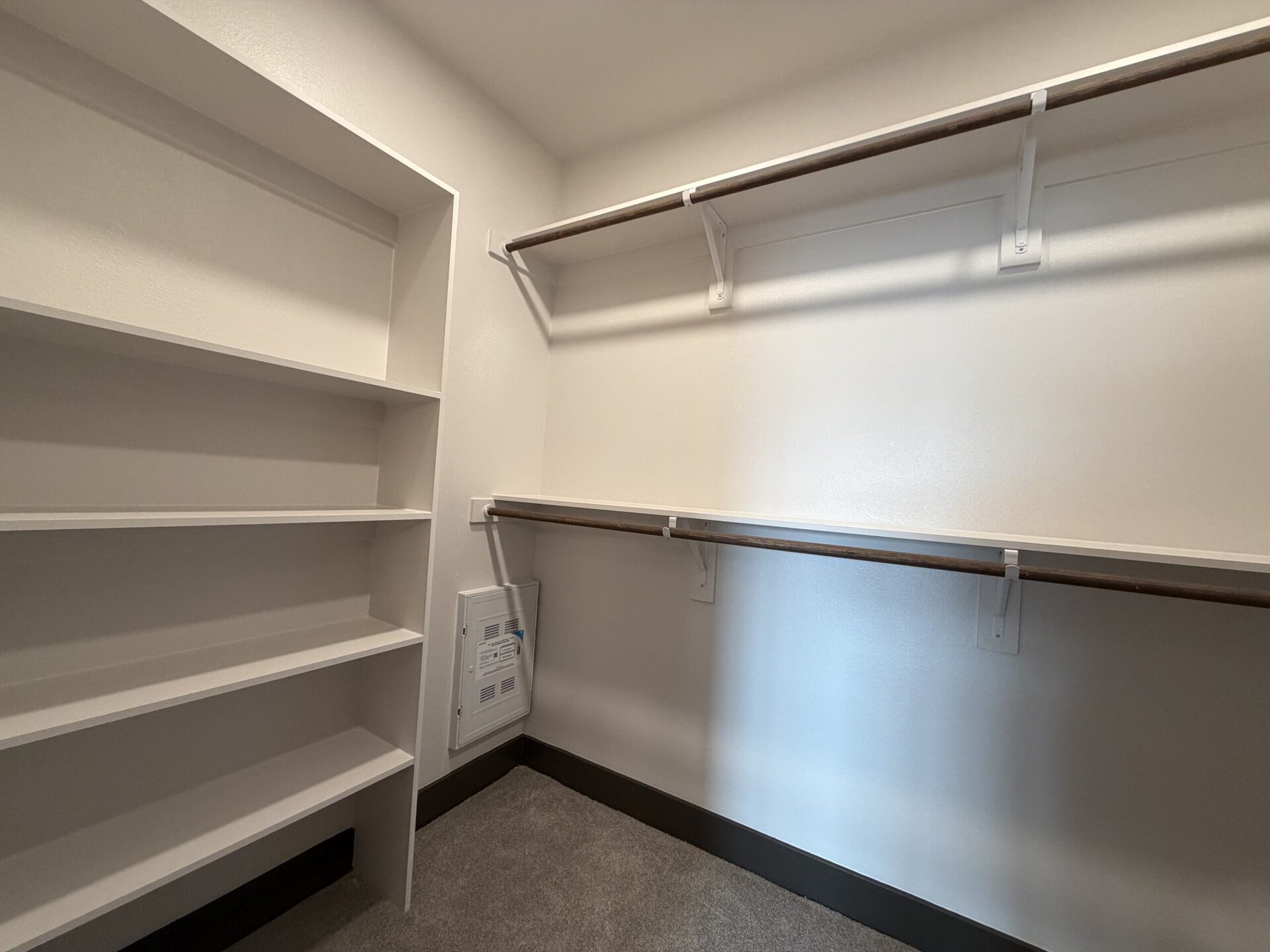 A spacious closet features two wooden hanging rods supported by white brackets, positioned against a light-colored wall. To the left, there is a white shelving unit with multiple open shelves, providing ample storage space. The floor is carpeted in a neutral tone, and a small electrical panel is mounted on the wall near the bottom right corner. The overall design is clean and minimalist.