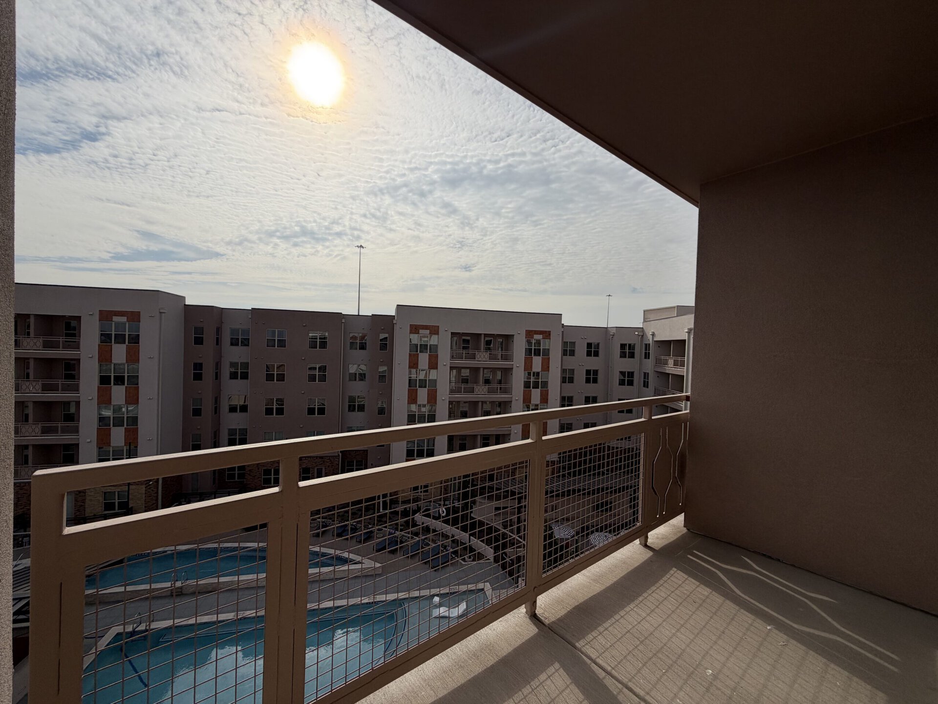 A balcony view overlooks a courtyard with several multi-story apartment buildings. The buildings feature a mix of light and dark facades, with some having decorative elements. A swimming pool is visible below, surrounded by lounge chairs. The sky is partly cloudy, with the sun shining brightly in the upper right corner. The balcony railing is a light color with a grid pattern.