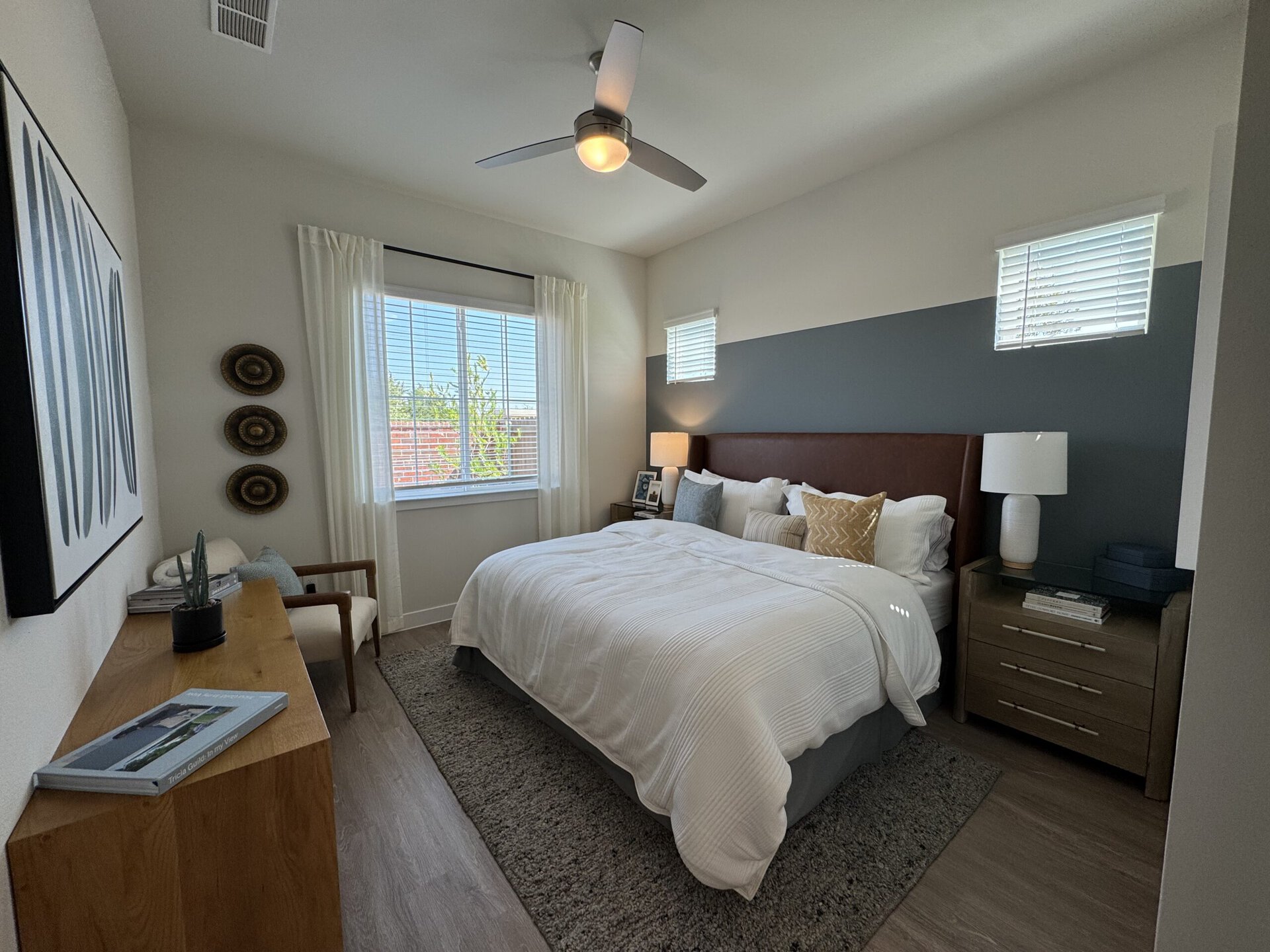 A cozy bedroom features a large bed with a white quilt and decorative pillows, including a gold accent pillow. The headboard is upholstered in brown leather. To the side, a nightstand holds a lamp and a framed photo. A wooden dresser with three drawers is positioned nearby. Natural light streams in through a large window adorned with sheer curtains, revealing greenery outside. On the opposite wall, three circular decorative pieces are displayed, and a piece of abstract art hangs above a wooden console table with books and a small potted plant. The flooring is light wood, and a soft gray rug lies beneath the bed.