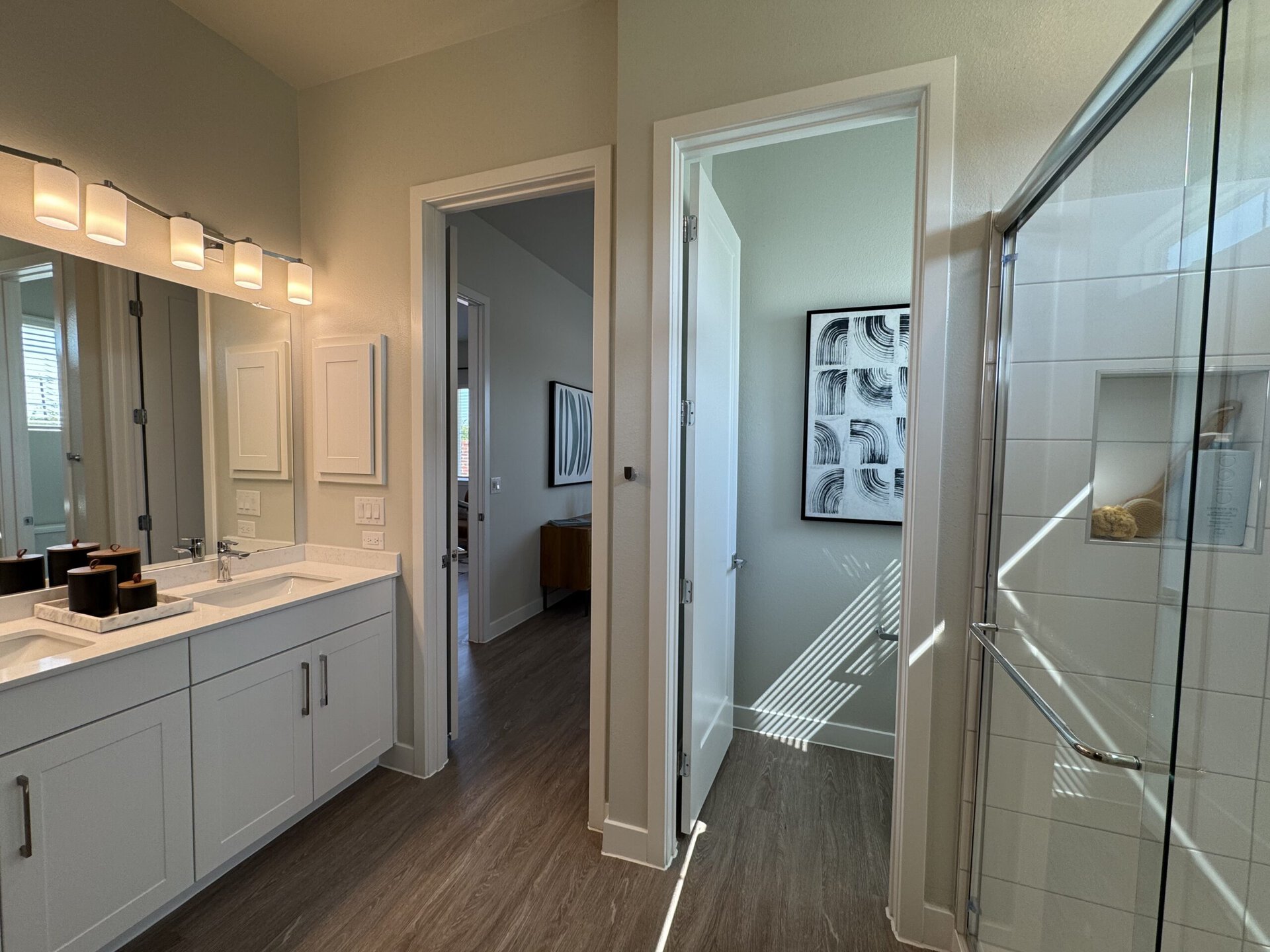 A modern bathroom features a double vanity with a white countertop and sleek cabinetry. Above the vanity, there are five cylindrical light fixtures providing illumination. A large mirror reflects the space. To the right, a glass shower enclosure showcases a tiled interior with a built-in shelf displaying bath products. Adjacent to the shower, there are two doors leading to separate rooms, one visible with a piece of abstract wall art and natural light coming through a window. The flooring is a warm wood tone, enhancing the contemporary aesthetic.