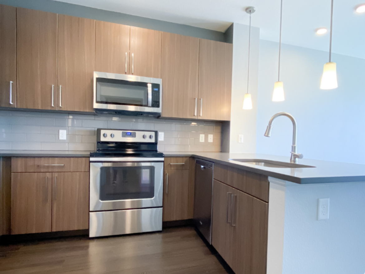 A modern kitchen featuring wooden cabinetry with a sleek finish, stainless steel appliances including a microwave and oven, and a dark countertop. The backsplash consists of light-colored tiles. There are two pendant lights hanging above the countertop, and a stainless steel sink with a curved faucet is positioned on the counter. The flooring is dark wood, contributing to the contemporary aesthetic.