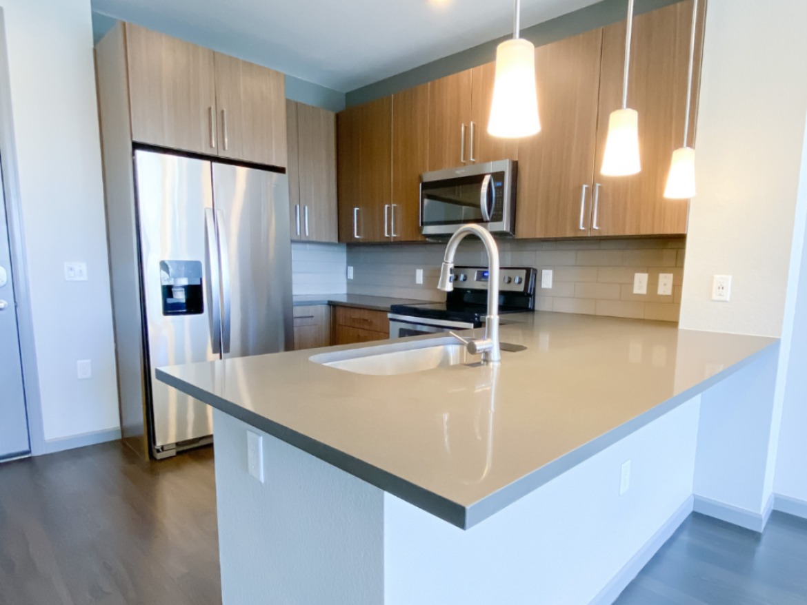 A modern kitchen featuring a large island with a smooth, light-colored countertop. The island includes a sink with a sleek faucet. Behind the island, there are wooden cabinets and stainless steel appliances, including a refrigerator and a microwave. The backsplash is made of light-colored tiles, and pendant lights hang above the island, providing illumination. The flooring is dark wood, creating a contrast with the lighter elements in the kitchen.