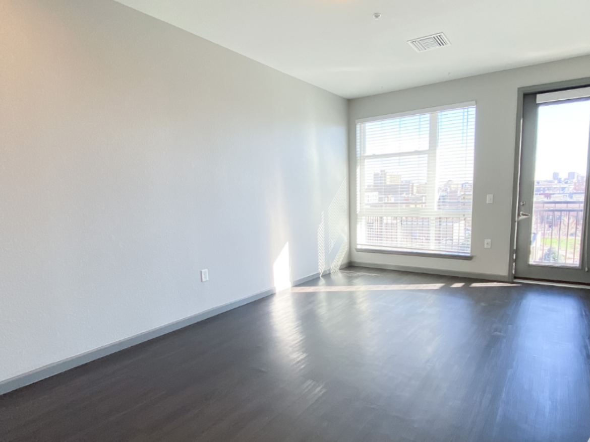 A spacious room with light gray walls and dark hardwood flooring. Large windows allow natural light to fill the space, with blinds partially drawn. A door leads to a small balcony, offering a view of the outside. The overall atmosphere is bright and airy, with minimal furnishings.