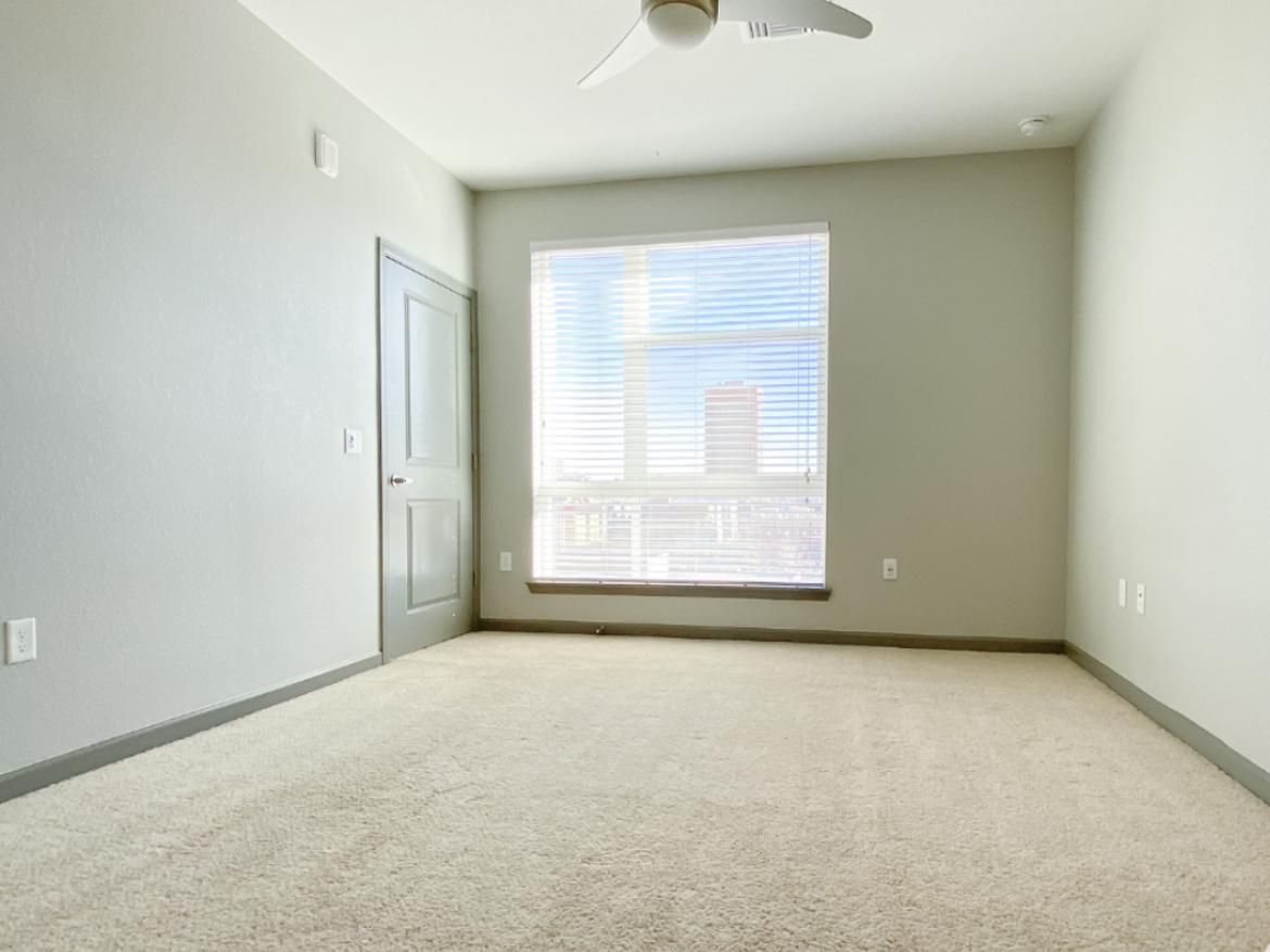 A spacious room with light gray walls and a light beige carpet. A window with blinds allows natural light to enter, revealing a view of a city skyline. A closed door is visible on the left side, and there are electrical outlets on the walls. The ceiling features a ceiling fan, adding to the room's functionality. The overall atmosphere is bright and airy, with a minimalist design.