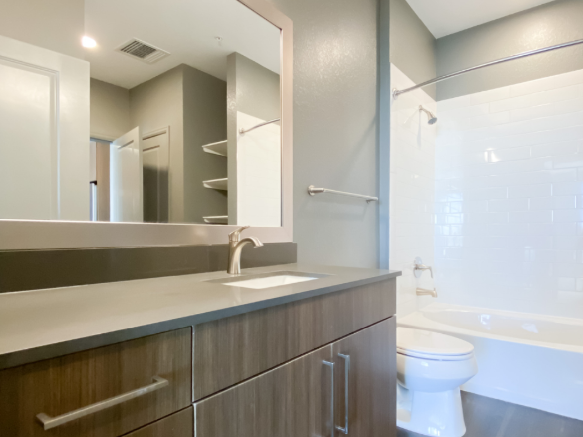 A modern bathroom features a sleek vanity with a gray countertop and a single sink. The cabinetry is a warm wood tone with minimalist handles. A large mirror above the sink reflects the space, which includes a shower area with white subway tiles and a bathtub. The walls are painted in a soft gray, and there are open shelves for storage. Natural light enters from an adjacent room, enhancing the bright and airy feel of the bathroom.