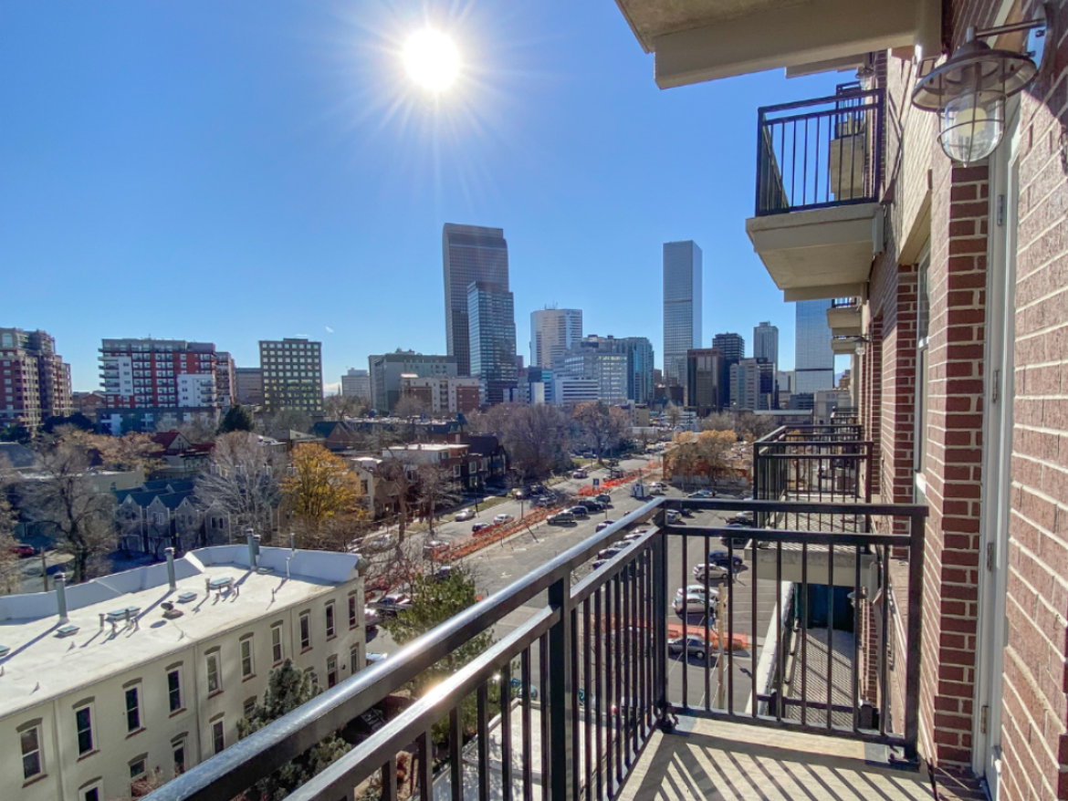 A view from a balcony overlooking a cityscape on a clear day. The sun shines brightly in the sky, casting a starburst effect. In the foreground, there are several balconies and a building with a white roof. Below, a street runs through the scene, lined with trees and various buildings, including modern skyscrapers and older structures. The skyline features a mix of architectural styles, with prominent tall buildings in the background.