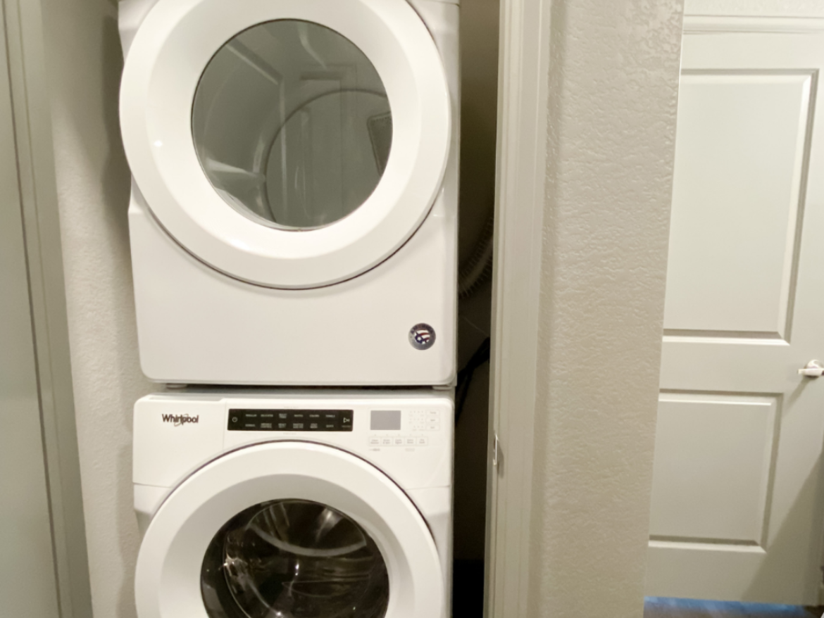 A stacked washer and dryer unit is positioned in a narrow space. The top unit is a dryer with a large circular glass door, while the bottom unit is a washer, also featuring a circular glass door. The appliances are white and have a control panel with buttons and a digital display on the front of the washer. The surrounding walls are painted in a light color, and a door is visible to the right, leading to another room.