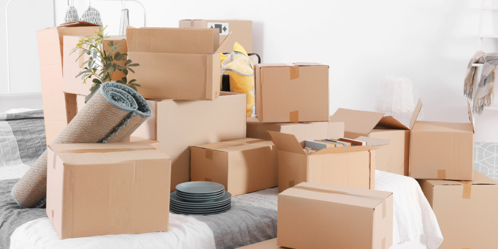 A cluttered room filled with various cardboard boxes of different sizes. Some boxes are stacked, while others are open, revealing items inside. A rolled-up rug is leaning against one of the boxes, and a small potted plant is visible among the boxes. There are also several gray plates stacked on the floor. The background features a bed with a gray and white blanket, adding to the sense of a recently moved-in space. Movers.
