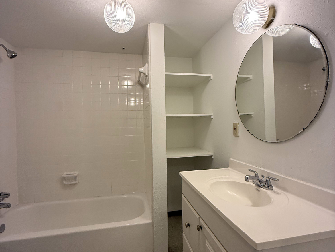 A small bathroom features a white tiled shower area with a bathtub. The showerhead is mounted on the wall, and there is a built-in shelf nearby. A circular mirror hangs above a white vanity with a single sink and chrome faucet. The vanity has two cabinet doors below. Two glass pendant lights illuminate the space, and there are additional open shelves on the wall for storage. The overall color scheme is bright and neutral.